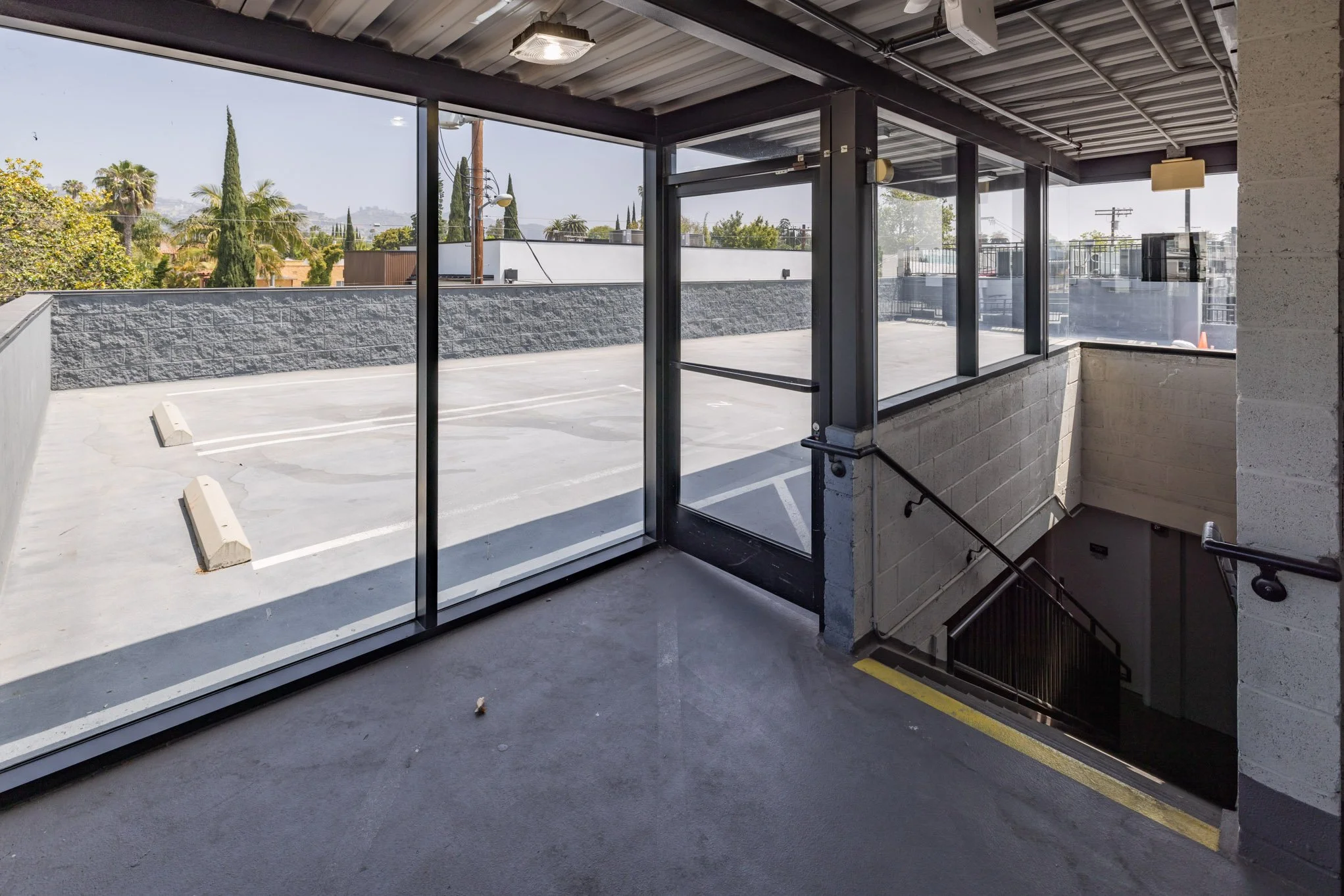 View of an outdoor parking lot with empty spaces, concrete parking barriers, and a surrounding gray brick wall, seen through a large glass enclosed stairwell lobby area with sliding glass doors inside a building.