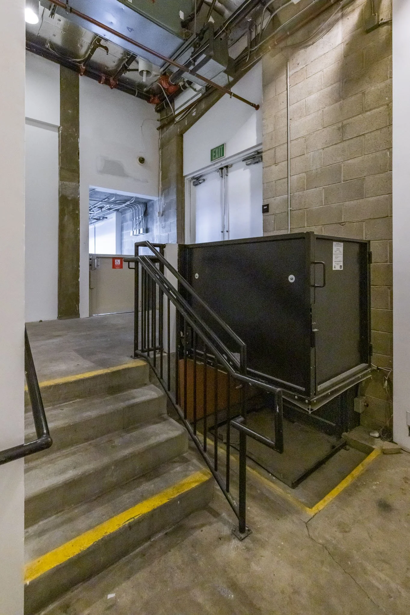 Emergency stairwell with black elevator lift, concrete stairs, metal railing, brick wall, and an exit door.