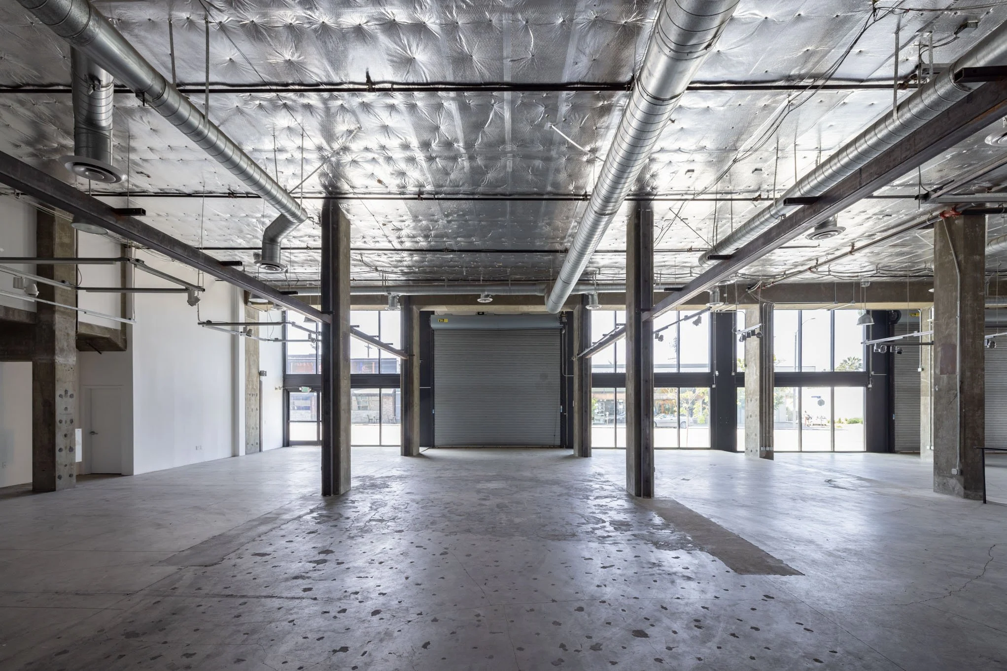 Empty commercial or industrial space with concrete floors, exposed ductwork and metal support columns, large windows, and a partially open metal roll-up door.