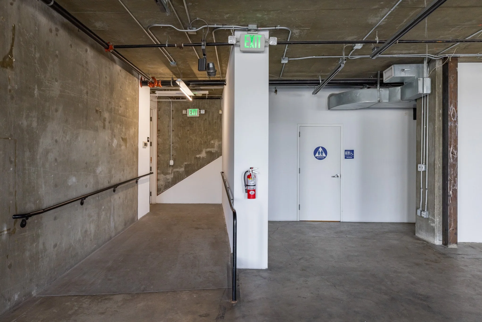 Underground parking garage corridor with concrete walls and floors, including an exit sign, fire extinguisher, and restrooms.