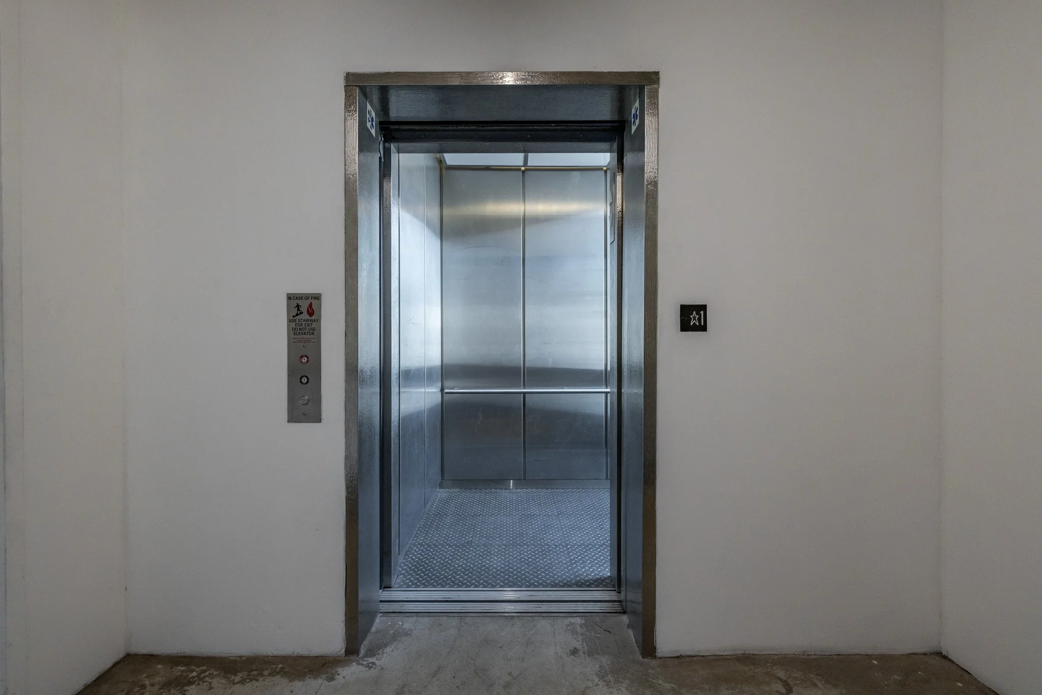 Elevator with open doors showing a metallic interior, wall on either side, and a floor with textured metal surface.