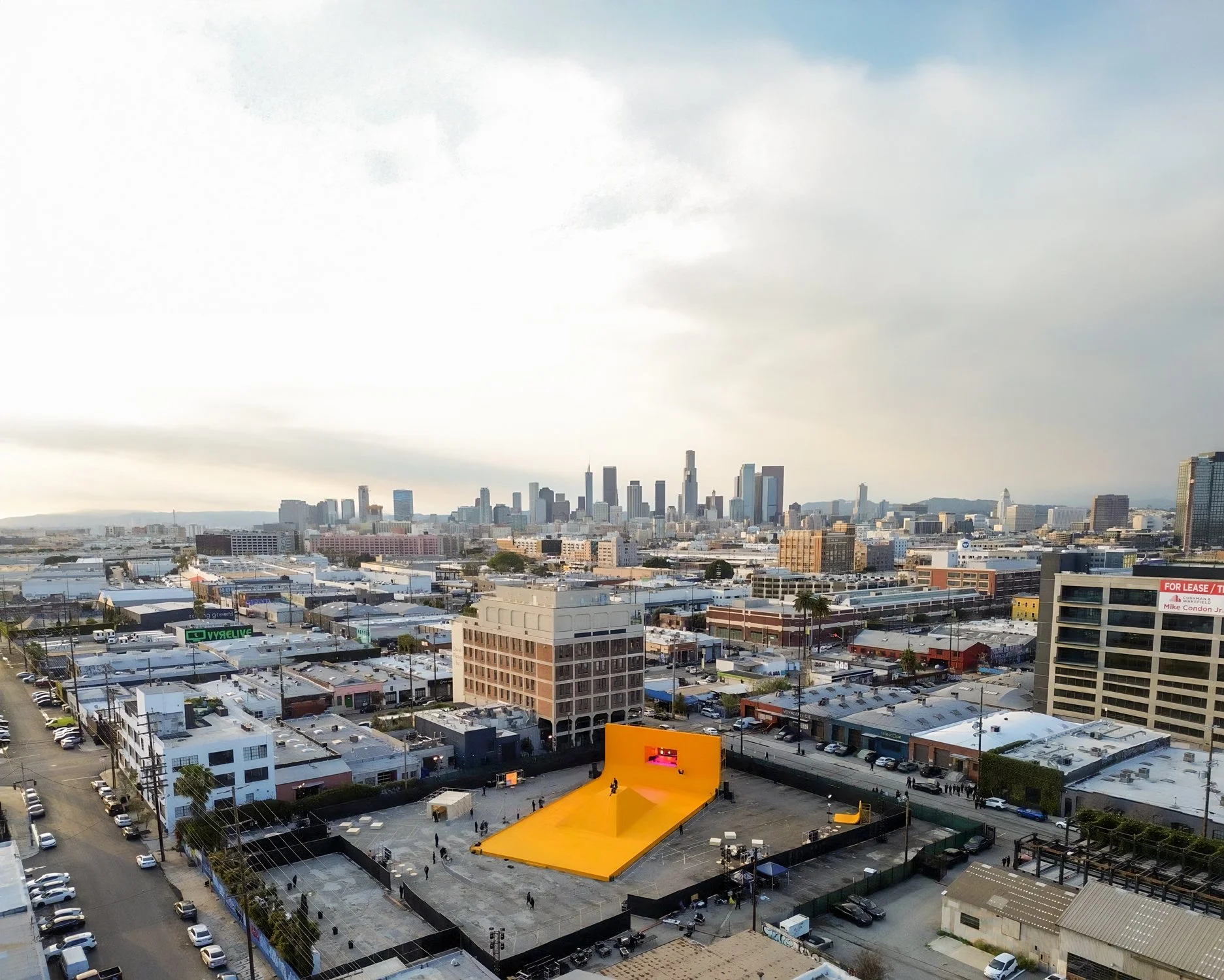 Aerial view of a city skyline with a yellow stage outdoors in the foreground, and several buildings and parking lots around it.