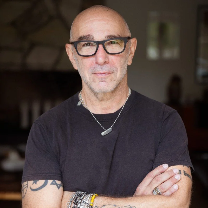 A man with a shaved head, wearing black glasses, a black t-shirt, and jewelry, standing indoors with his arms crossed and smiling slightly.