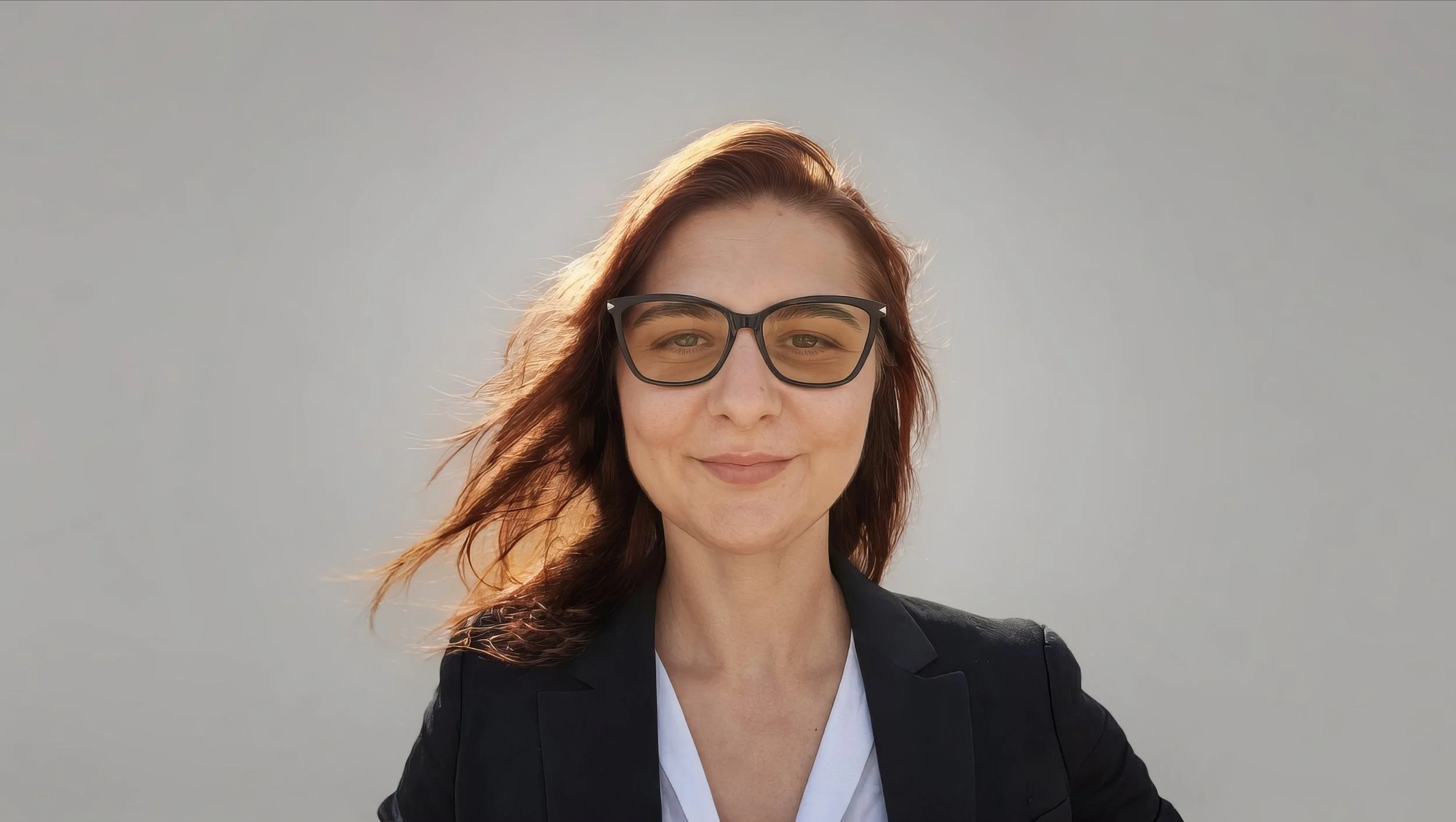 A young woman with long, reddish-brown hair, wearing glasses and a black blazer over a white top, is smiling against a plain grey background.