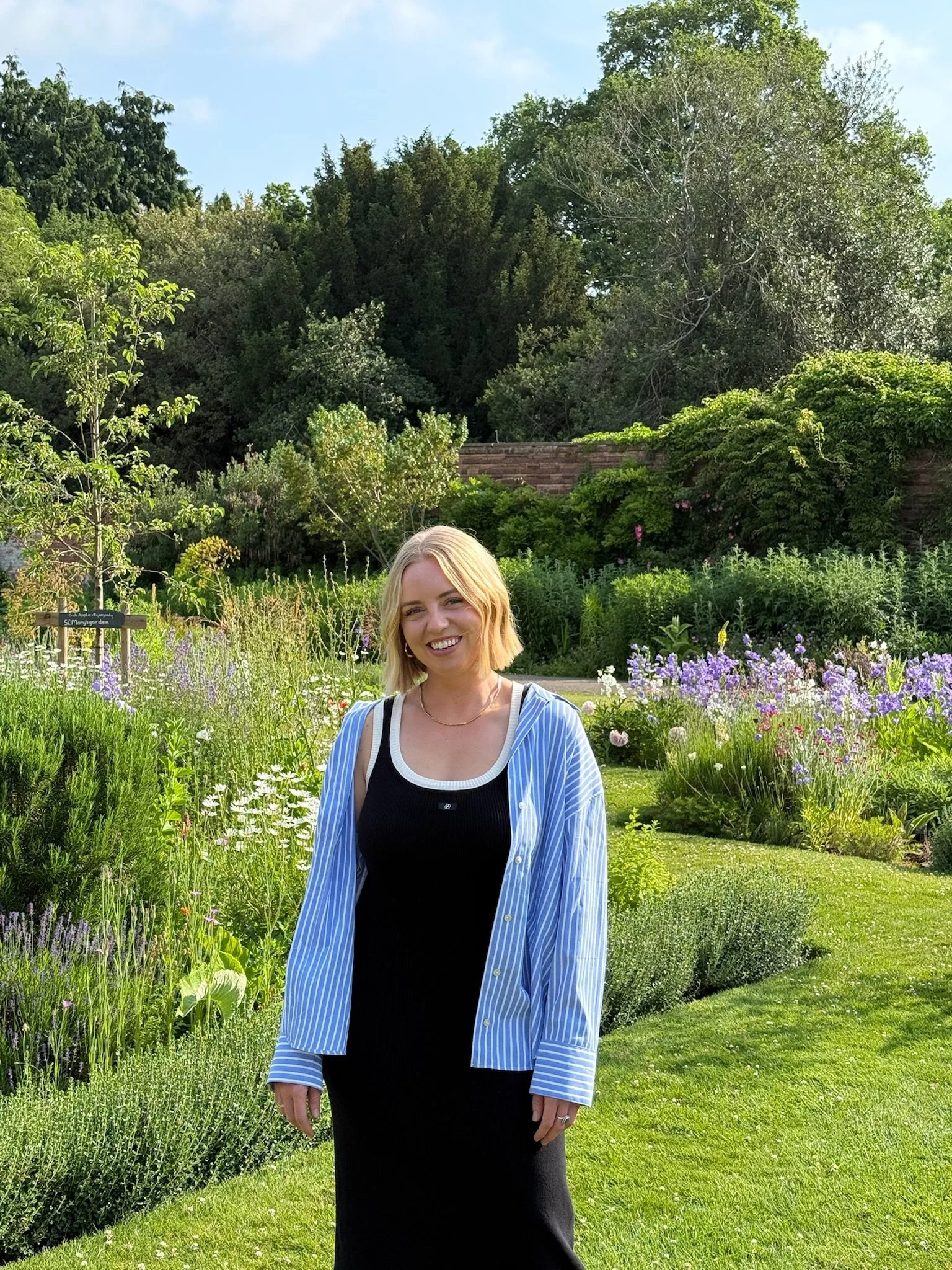 A woman standing in a garden with colorful flowers and green trees in the background, smiling at the camera.