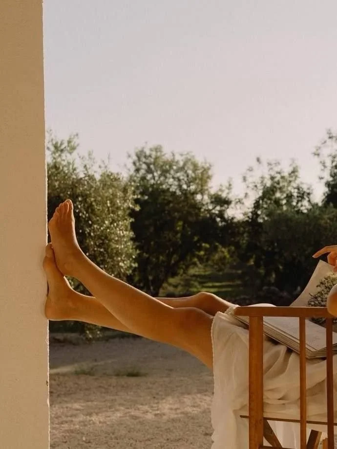 Person relaxing outdoors, lying on a chair with legs extended and feet resting against a wall, holding a book.