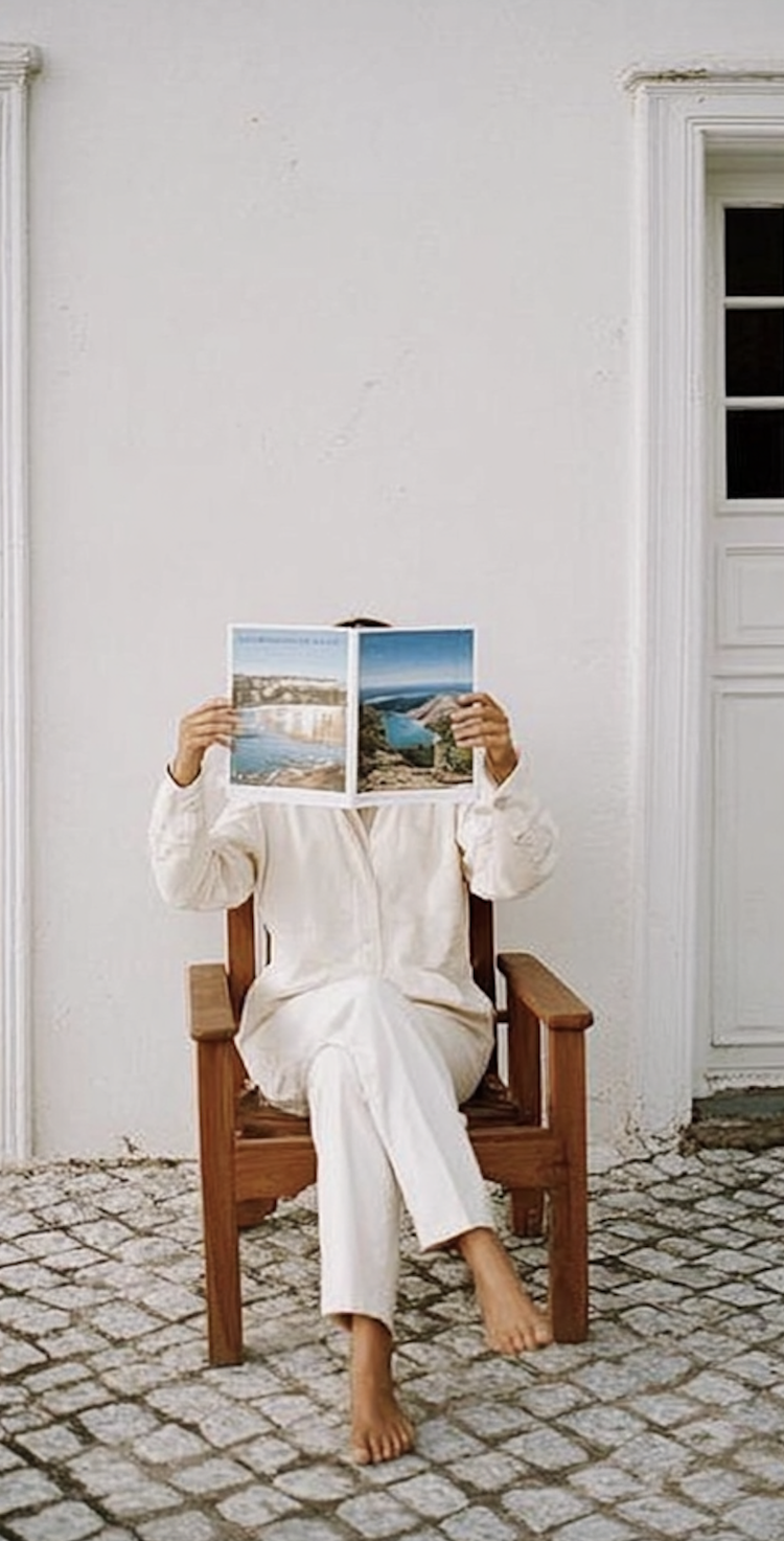 Person in white sitting on a wooden chair reading a brochure outdoors