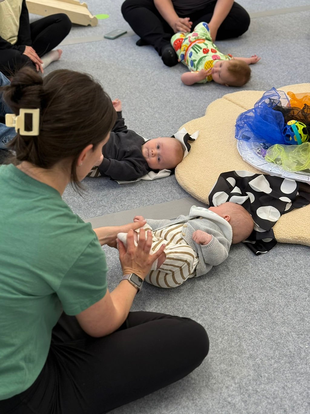 An adult woman with brown hair in a bun, wearing a green shirt, interacts with a baby lying on the floor. Other babies are also lying nearby, and there are soft mats and colorful toys around.