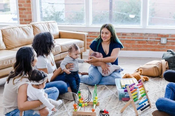 A woman with blue hair sitting on the floor playing with a group of children in a bright living room. There are toys and a plush bear on the floor, and a beige couch and large window in the background.