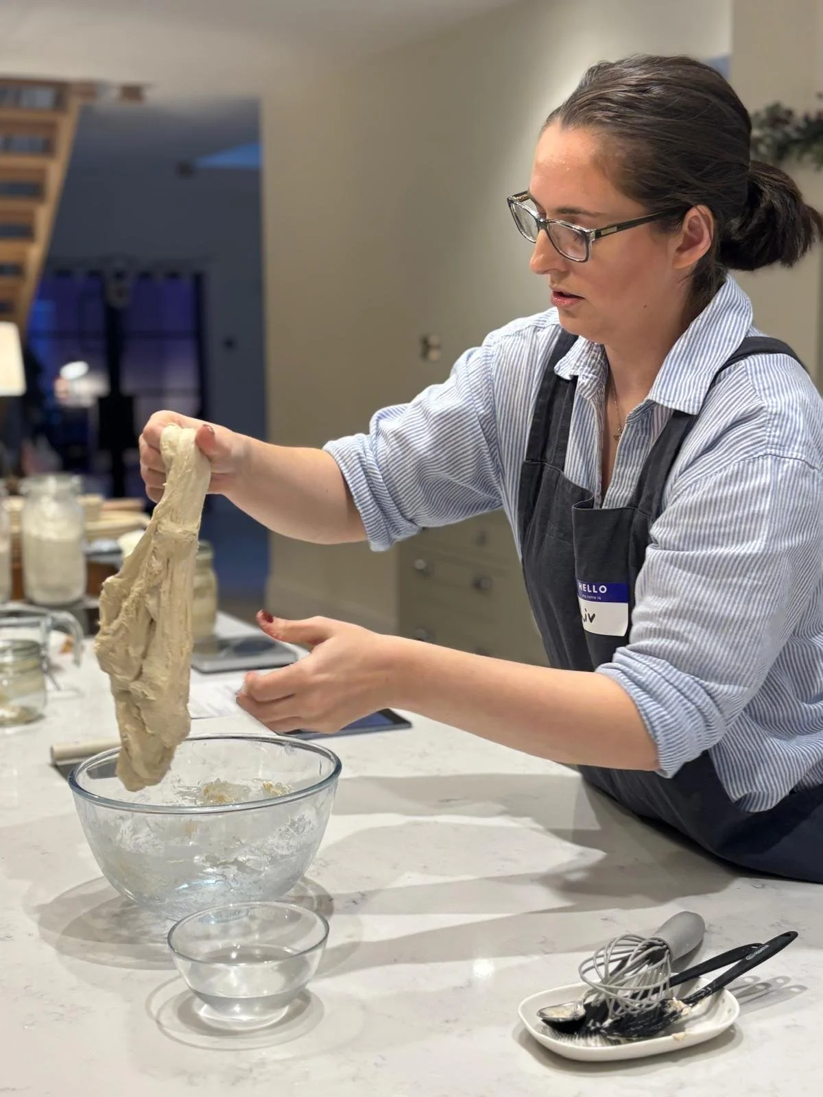 A woman with glasses, wearing a striped shirt and apron, is lifting dough from a glass mixing bowl in a kitchen.