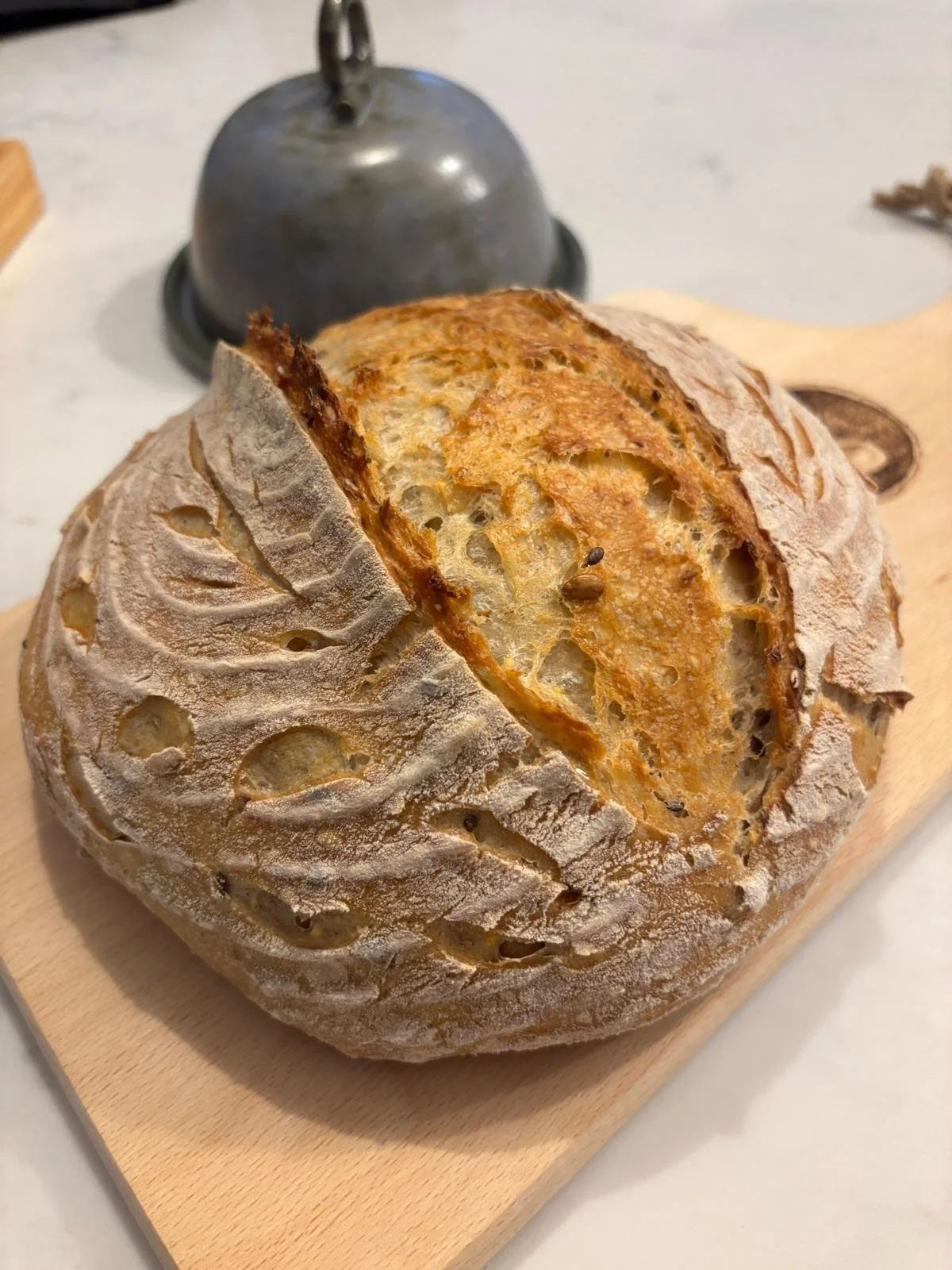 A round loaf of crusty bread with a scored top, resting on a wooden board with a metal bell and a marble surface in the background.