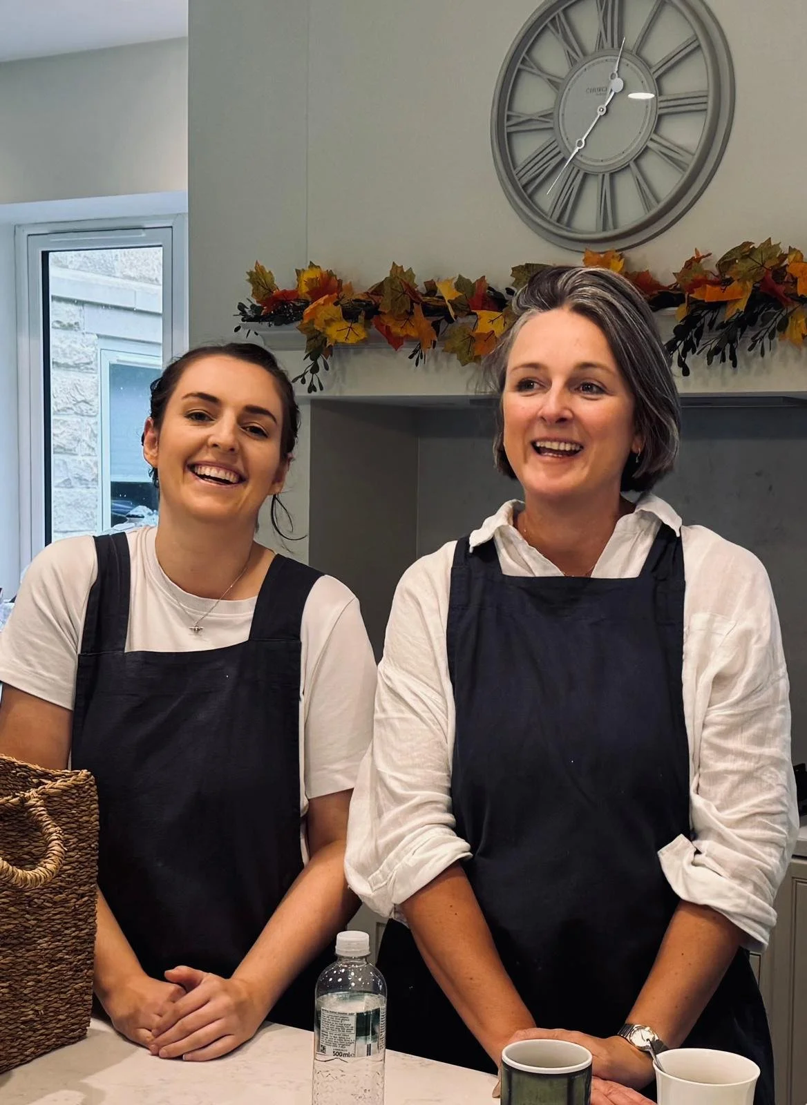 Two women smiling while standing in a kitchen, wearing black aprons, with a decorative autumn garland above them and a clock on the wall.