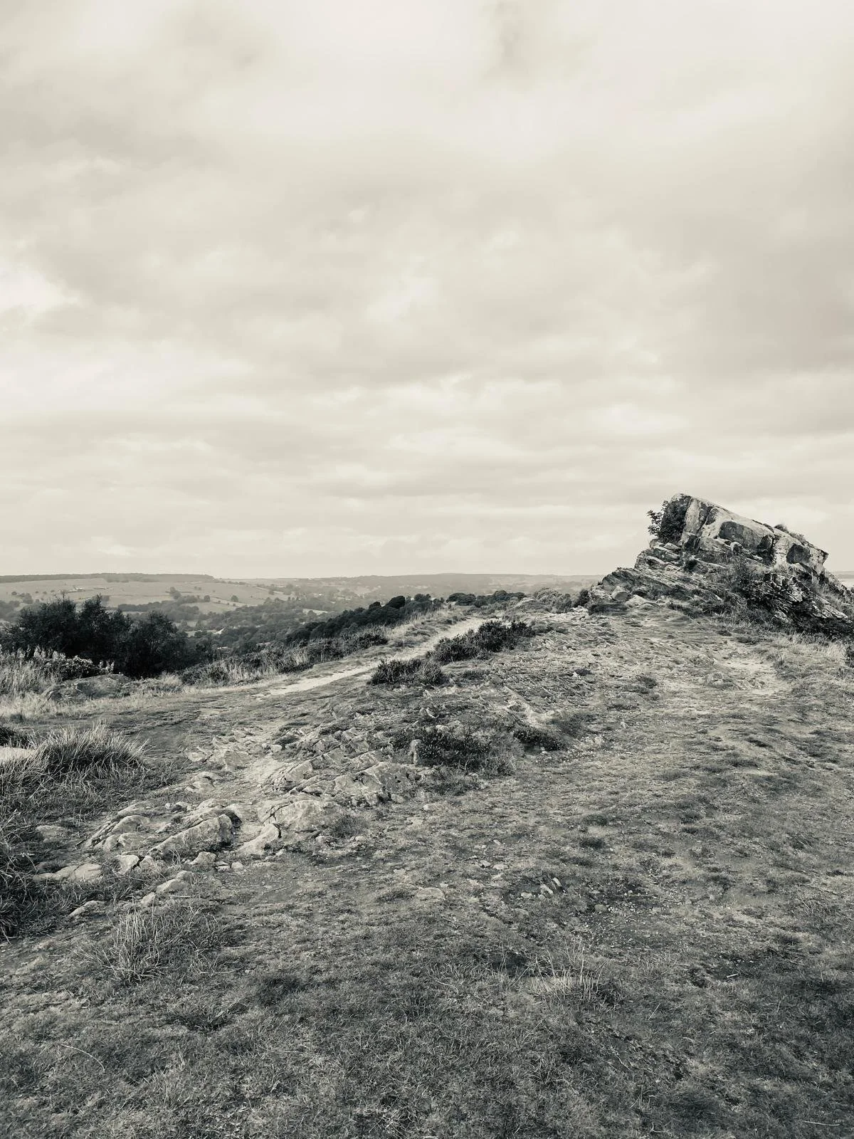 A rocky and grassy hilltop with a large tilted rock formation on the right, overlooking a landscape with hills and a cloudy sky.