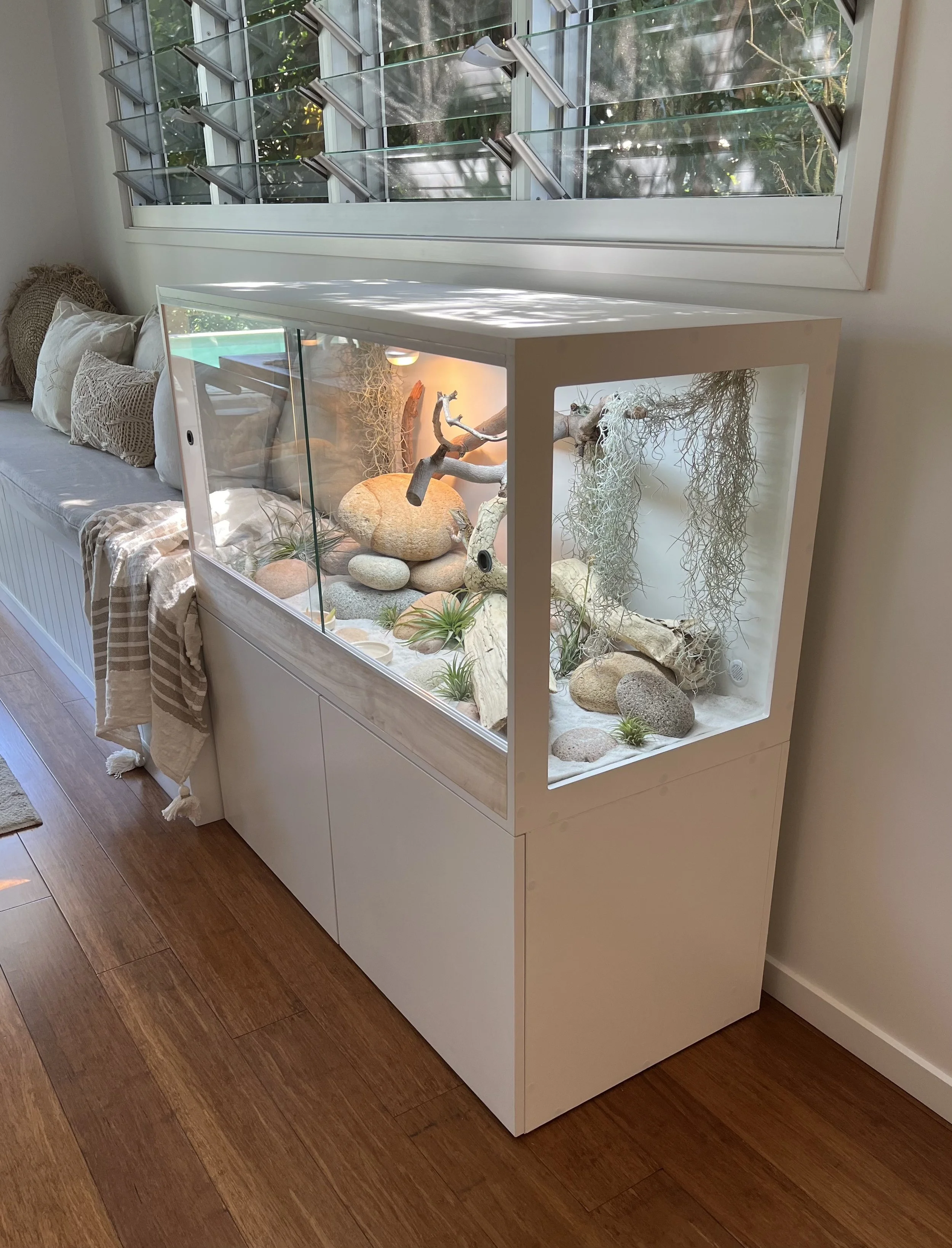 A modern terrarium with glass walls, containing rocks, driftwood, Spanish moss, and small plants, placed on a white cabinet next to a window.