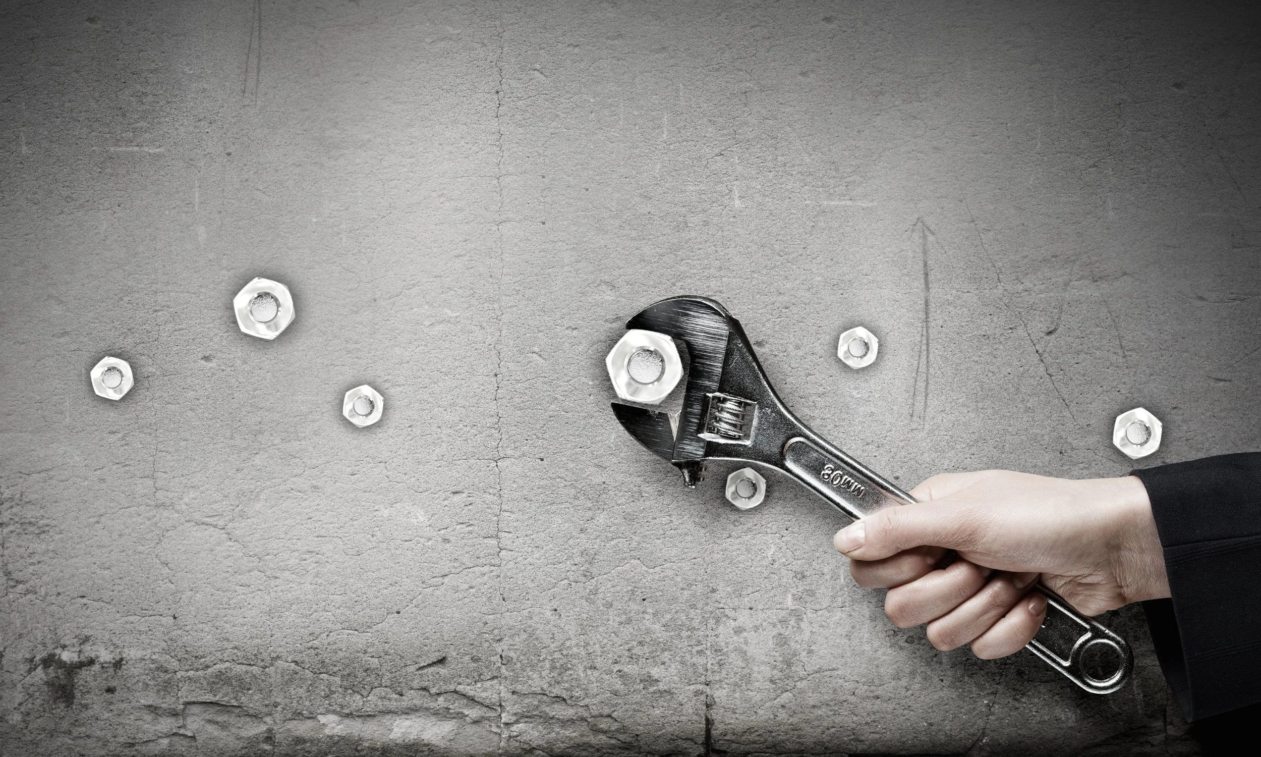 A person's hand holding a wrench on a concrete wall with several nuts attached to the wall, some in the process of being secured.