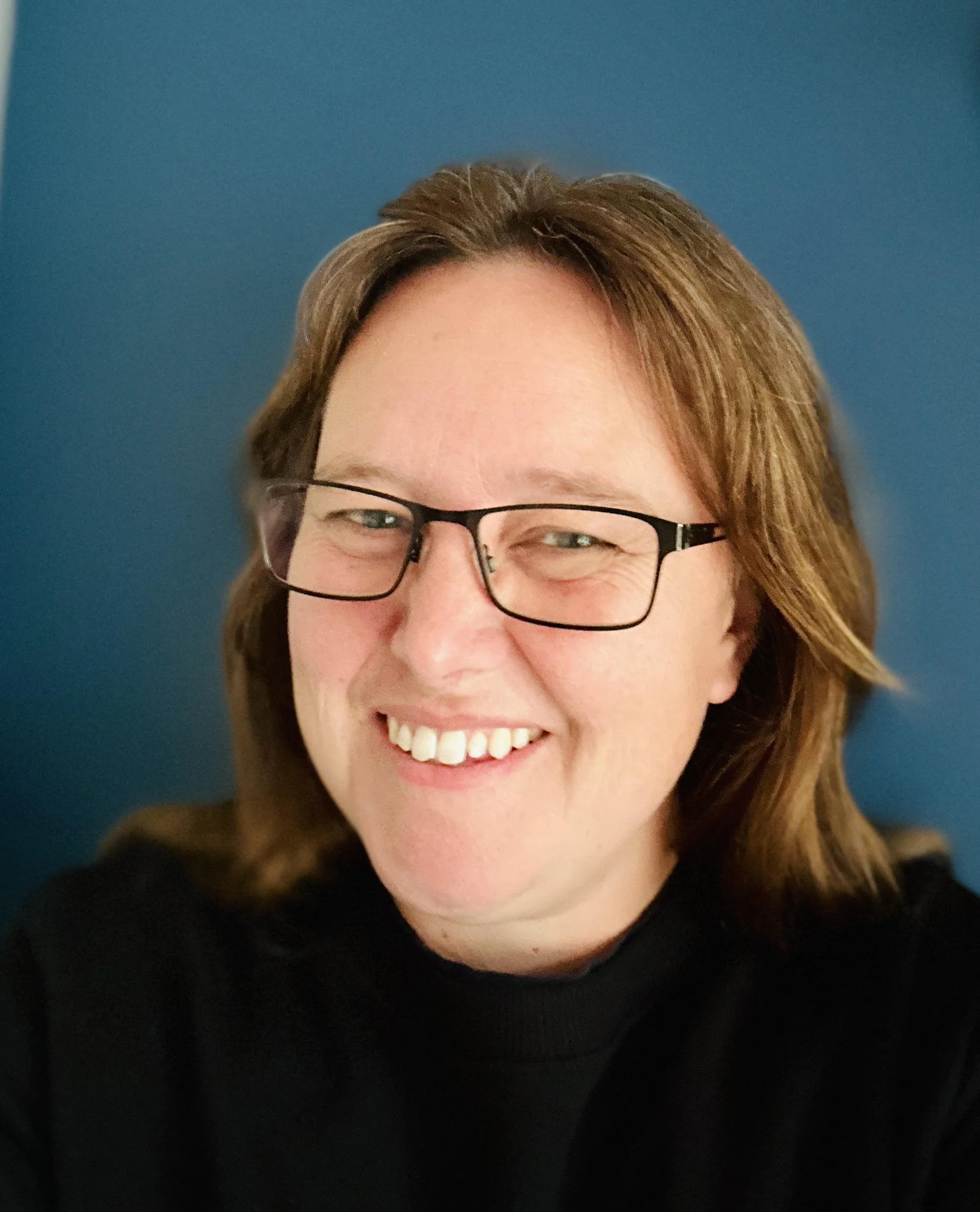 A person with shoulder-length brown hair, glasses, and a smile, wearing a black shirt, standing against a blue background.