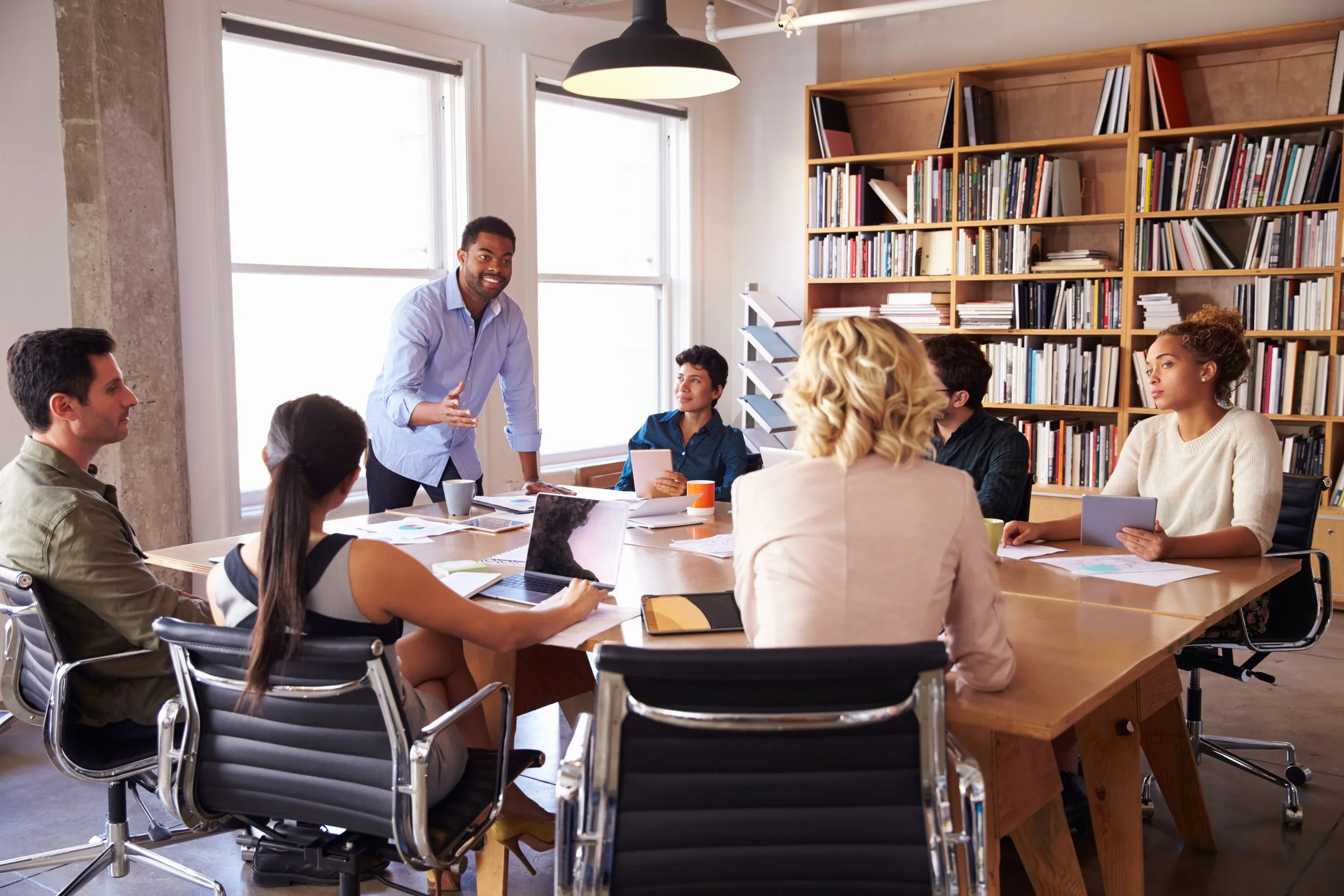 A diverse group of people in a meeting room listening to a man speaking, with shelves of books in the background.