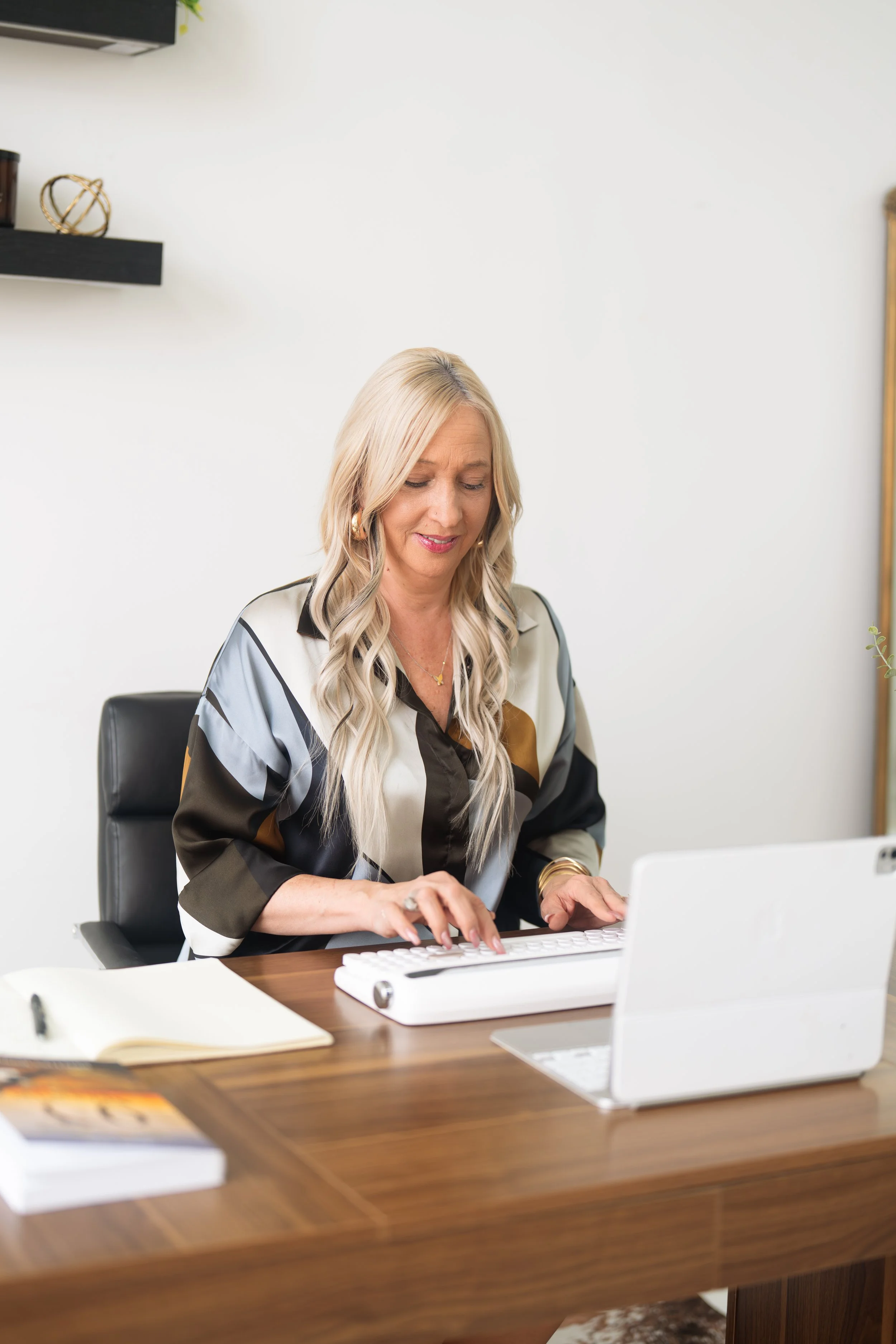 A woman with long blonde hair sitting at a wooden desk typing on a keyboard in a modern office setting.