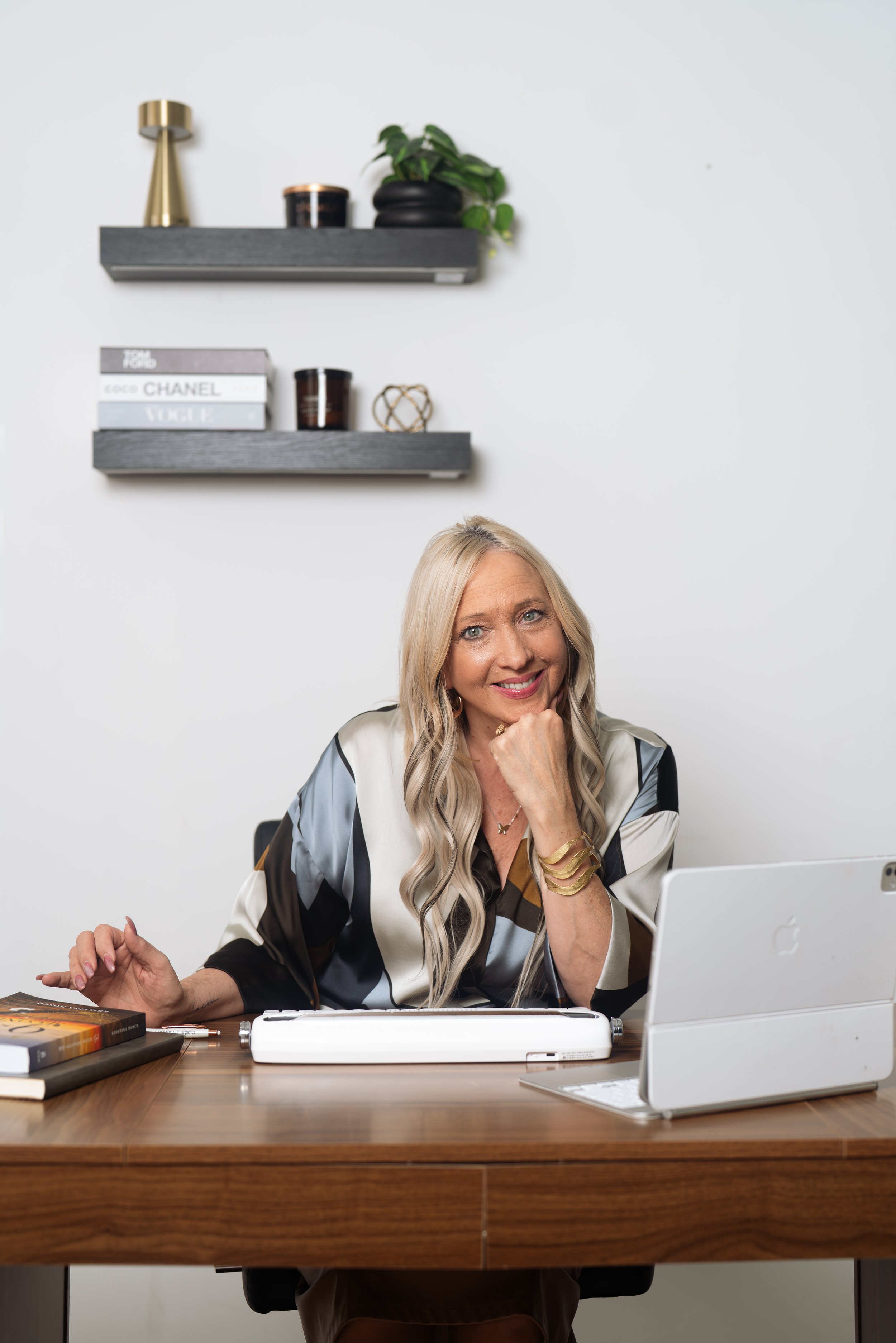 A woman sitting at a desk with a laptop, books, and a typewriter, smiling, with decorative shelves and books on the wall behind her.