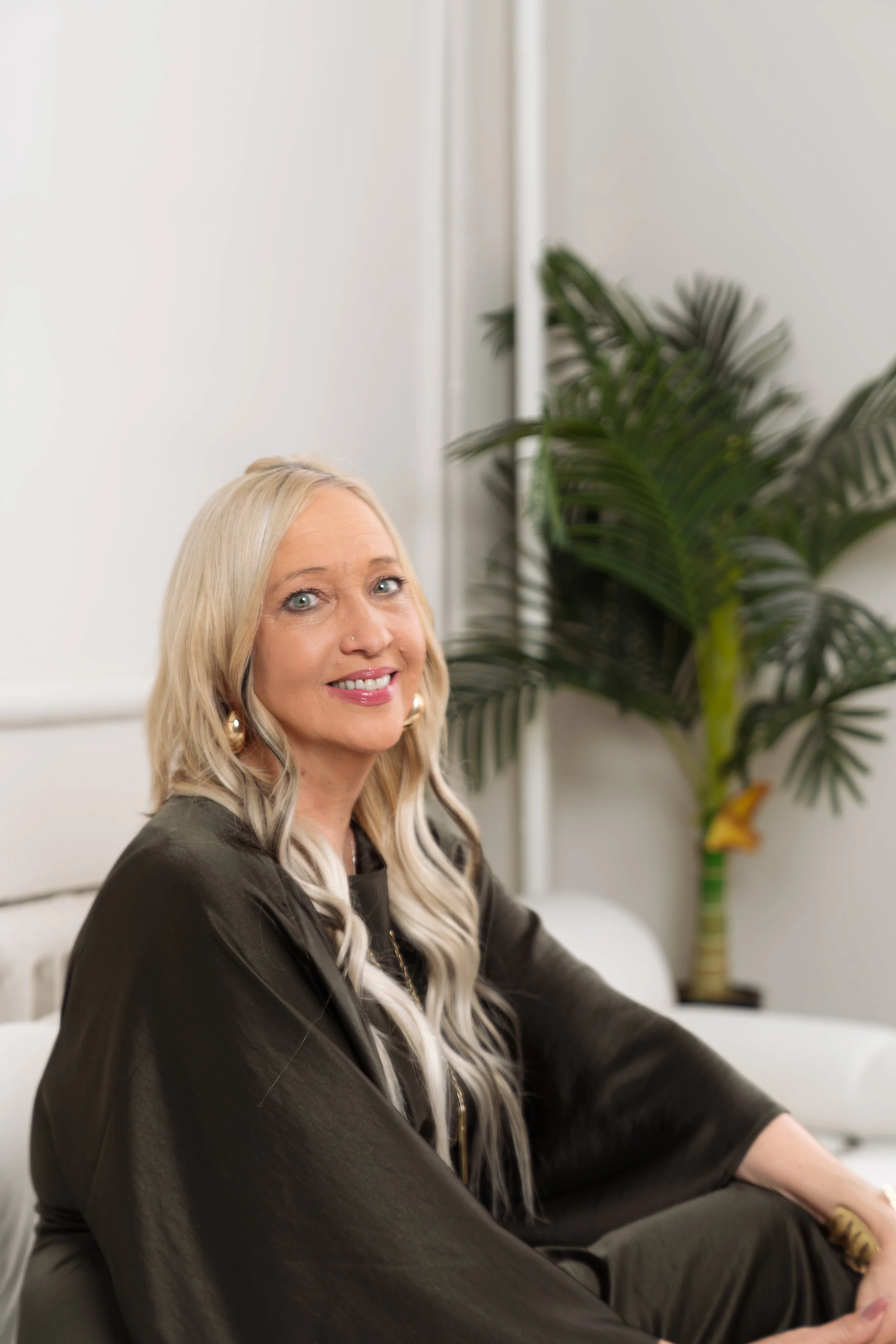 A woman with long blonde hair, wearing a dark jacket, sitting on a white couch in a room with white walls and a large potted plant in the background.