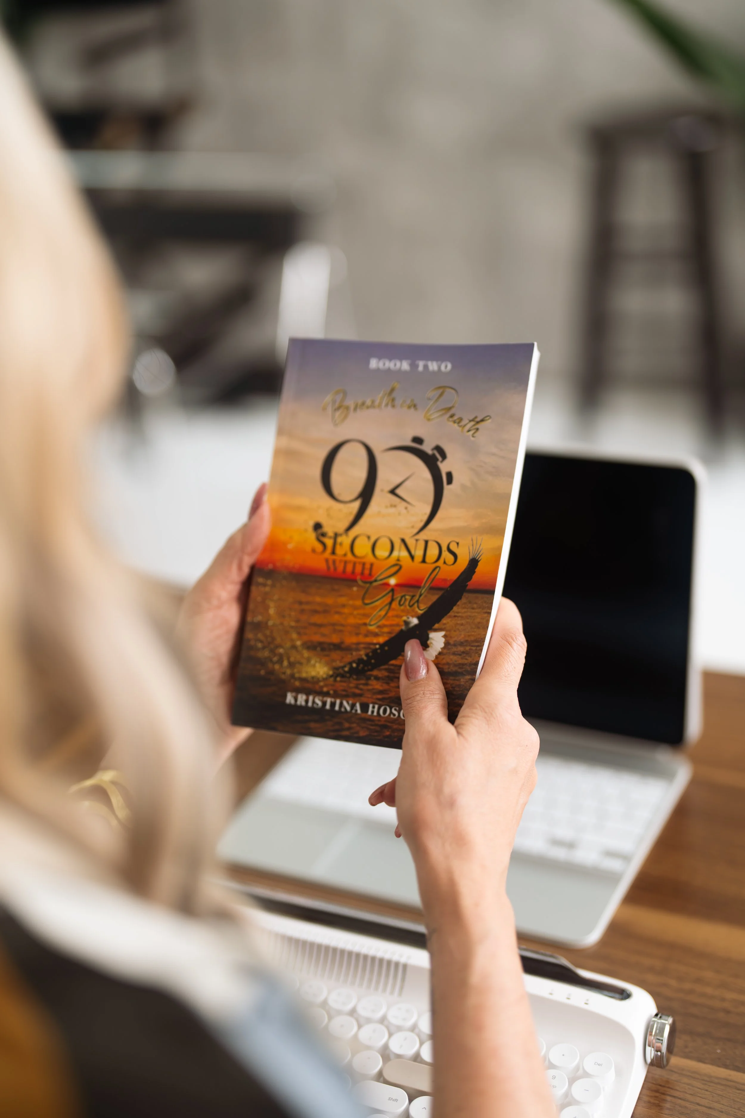 A woman holding a book titled '9 Seconds with God' while sitting at a desk with a laptop and a keyboard.