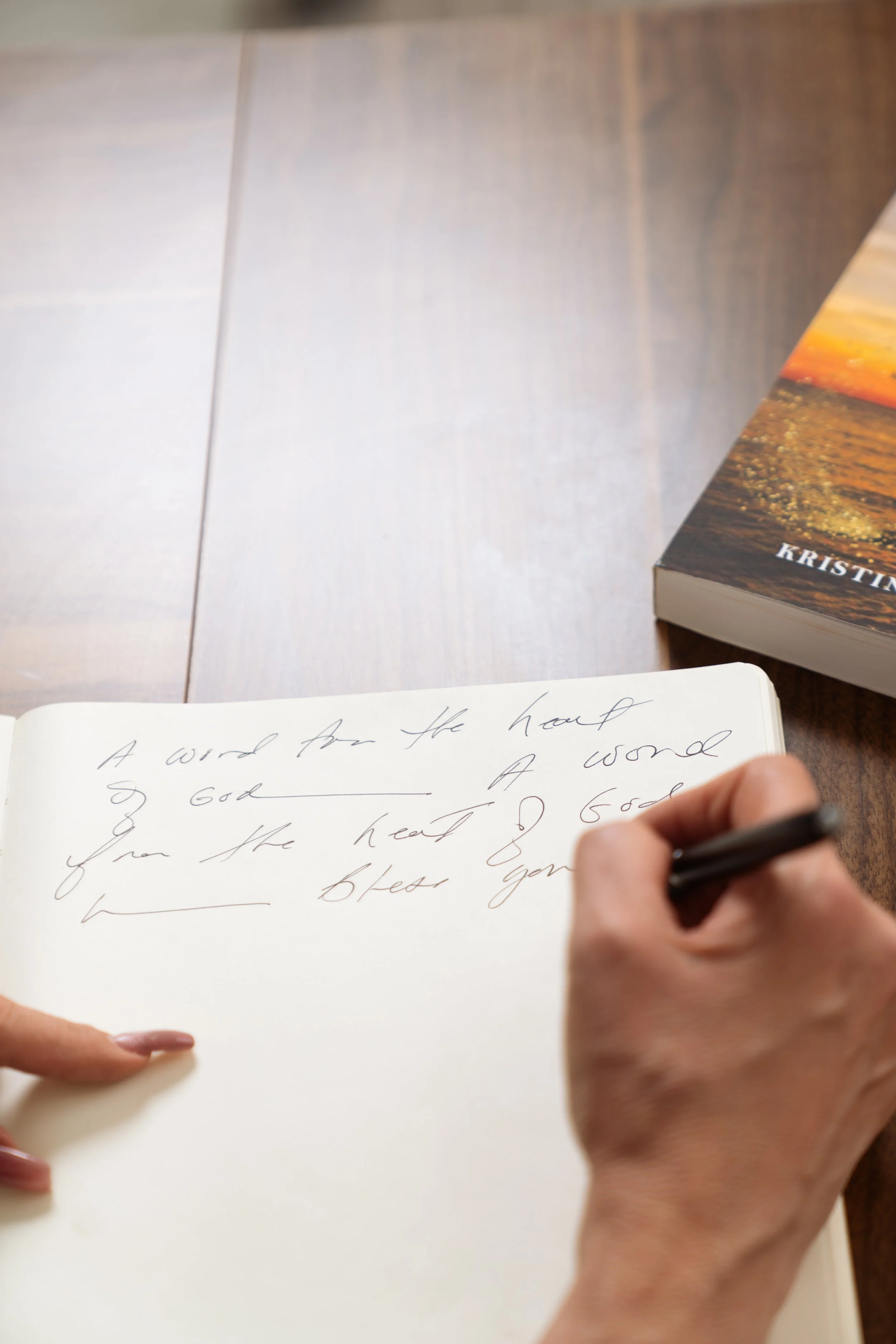 Handwriting in a notebook that reads, 'A word from the heart of God,' with a person writing with a black pen, and a paperback book titled 'Kristin' on a wooden table.
