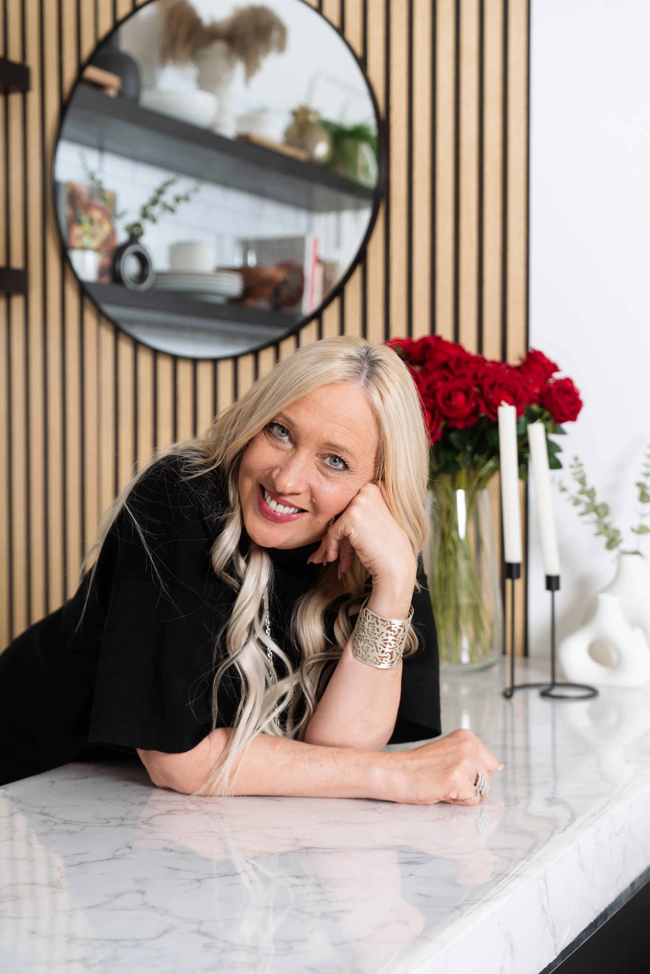 A smiling blonde woman with wavy hair lying on a white marble countertop with her head resting on her hand, in a modern interior with a bouquet of red roses and decorative candles in the background.