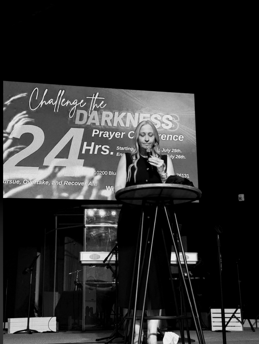 A woman standing at a podium speaking into a microphone during a prayer conference, with a large banner behind her that says 'Challenge the Darkness Prayer Conference 24 Hrs.'