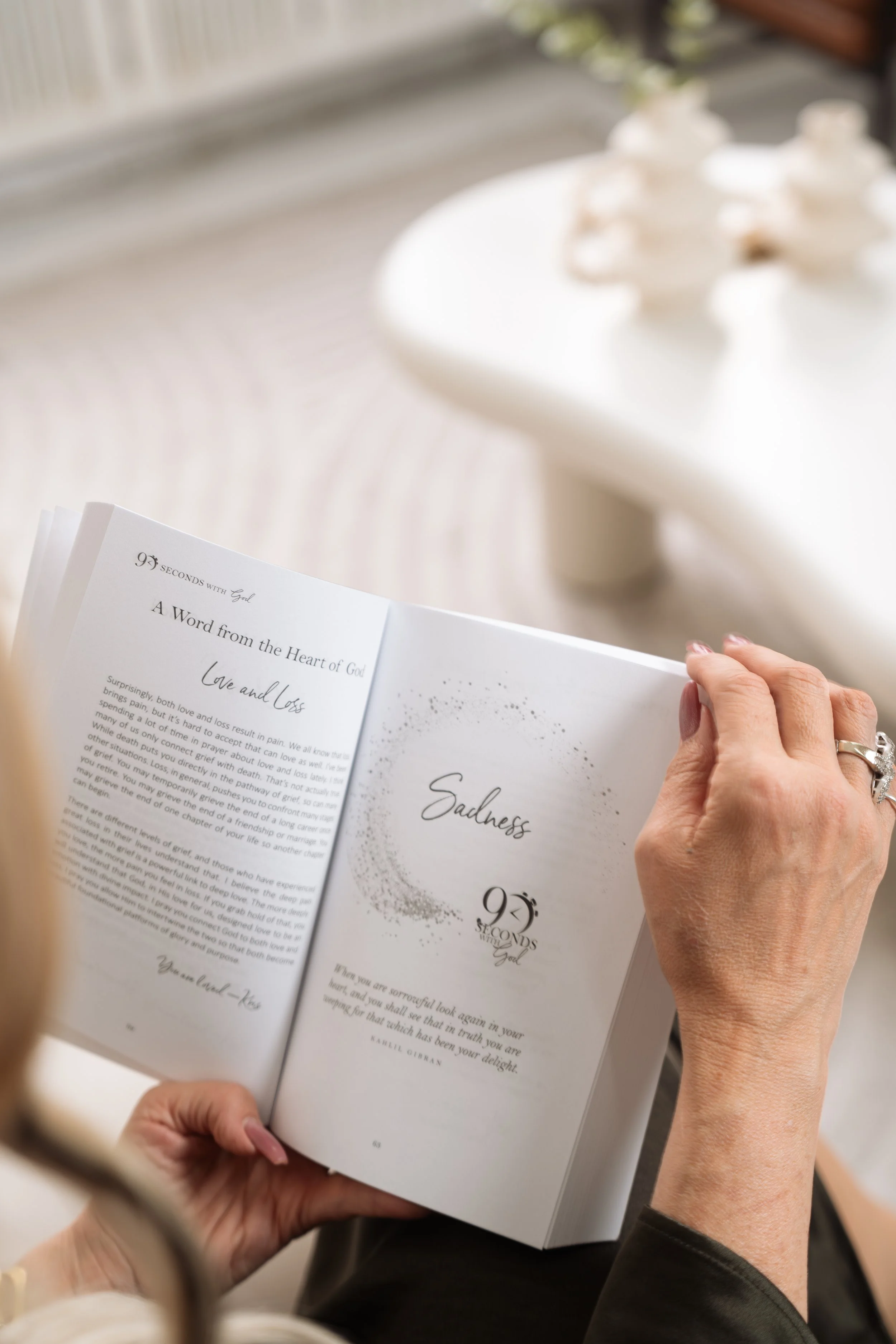 Person holding an open booklet with the word 'Sadness' on the page, beside a table with decorative items.