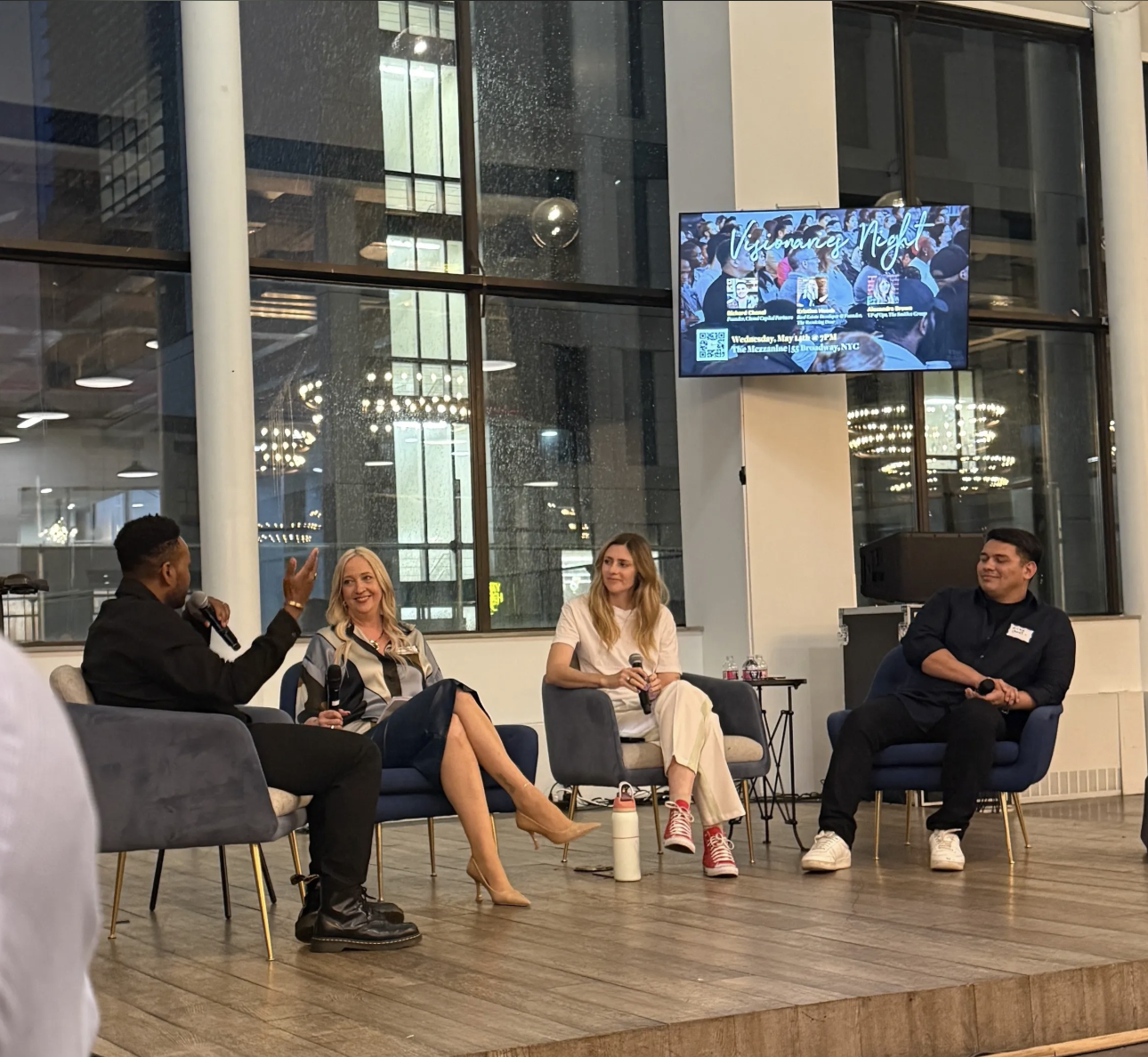 Panel discussion with four people seated on stage, two women and two men, with a large window behind them and a screen displaying event details, in a modern, well-lit room.