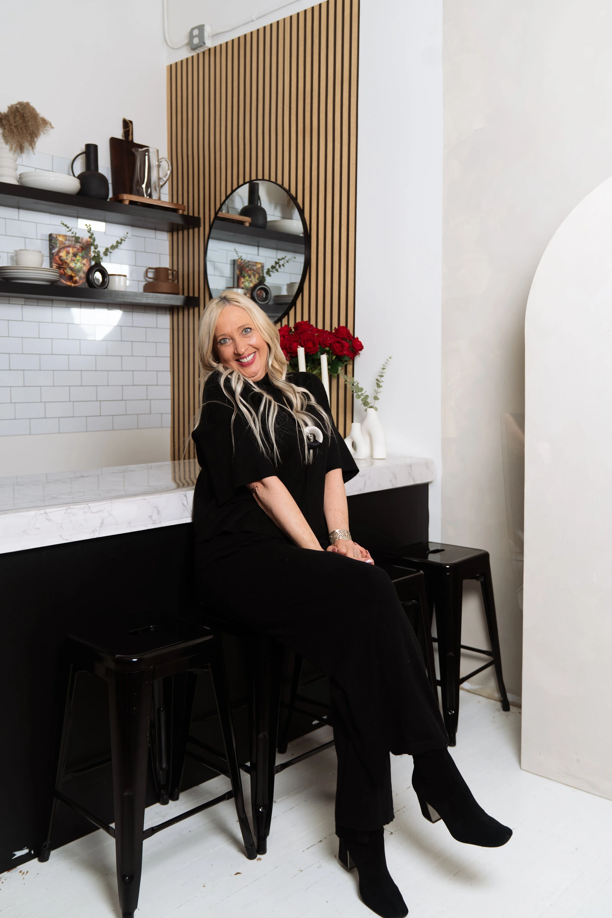 A woman with long blonde hair, wearing a black outfit and black high-heeled boots, sitting on a black stool in a modern kitchen with white subway tiles, wooden accents, and decorative items, smiling at the camera.