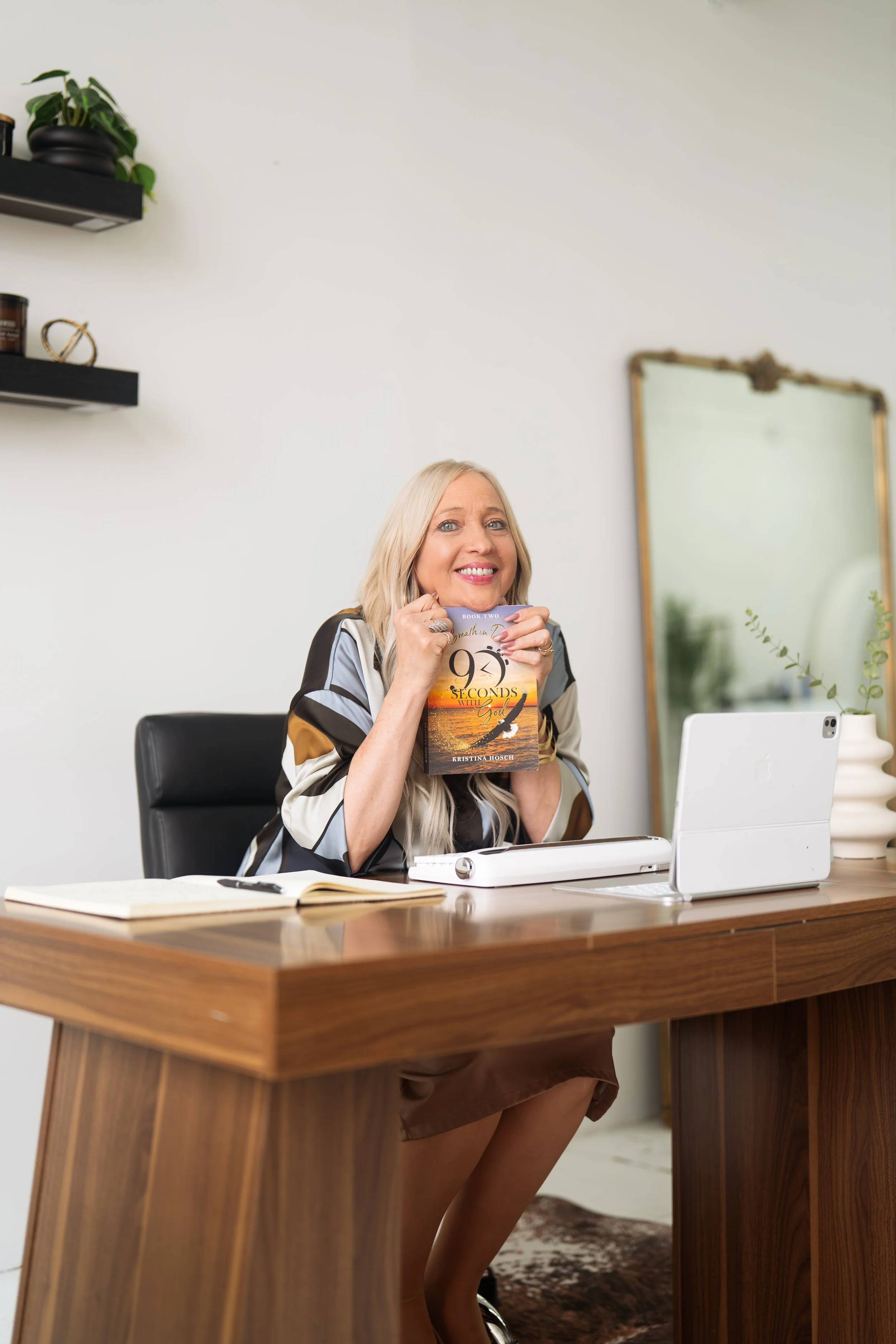 A woman sitting at a wooden desk smiling and holding a book titled '99 Seconds.' She is in a modern office with a mirror and shelves in the background, a notebook, and a tablet on the desk.