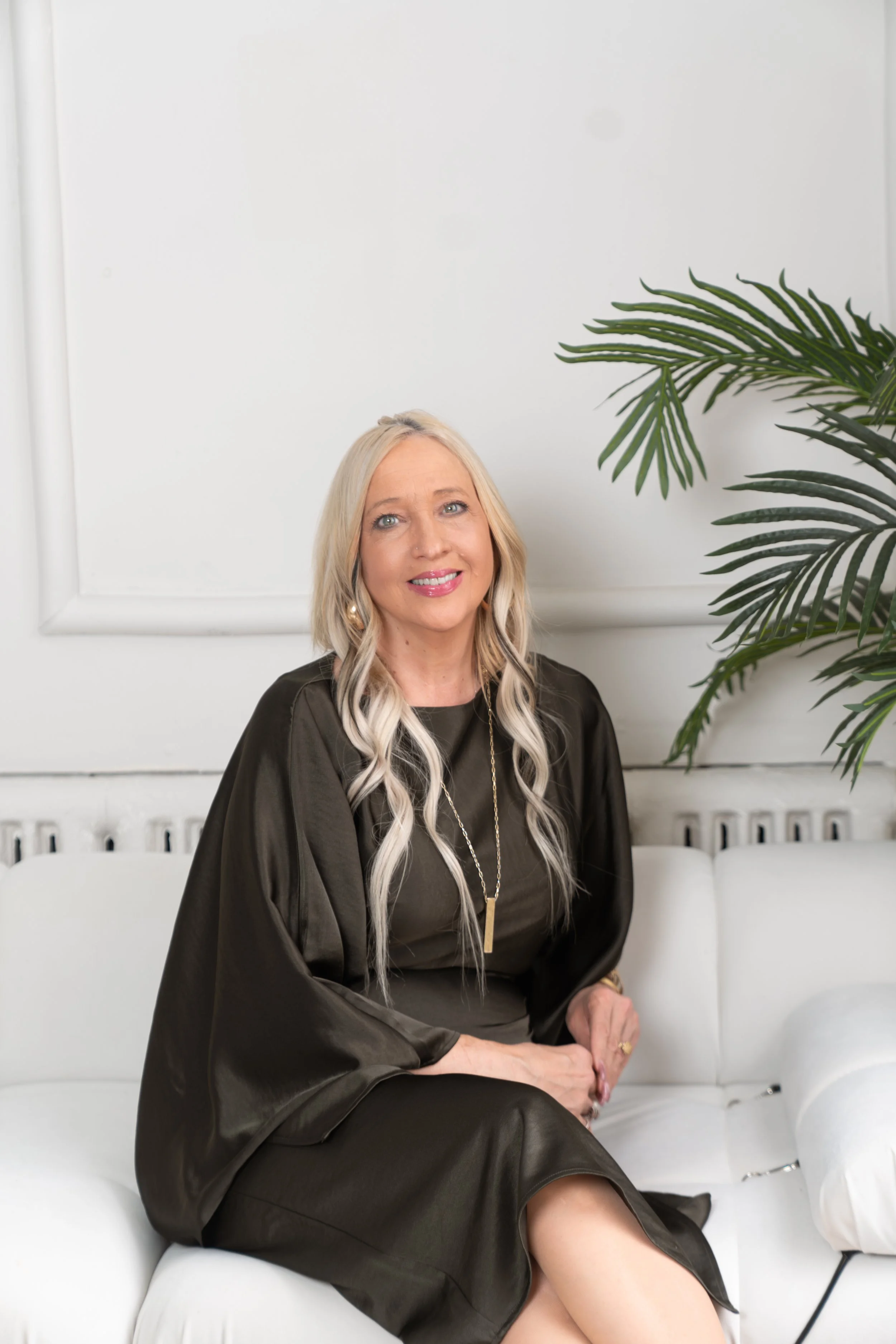 A woman with long blonde hair wearing a black dress with shiny fabric, sitting on a white couch in a room with white walls and a large green palm plant.