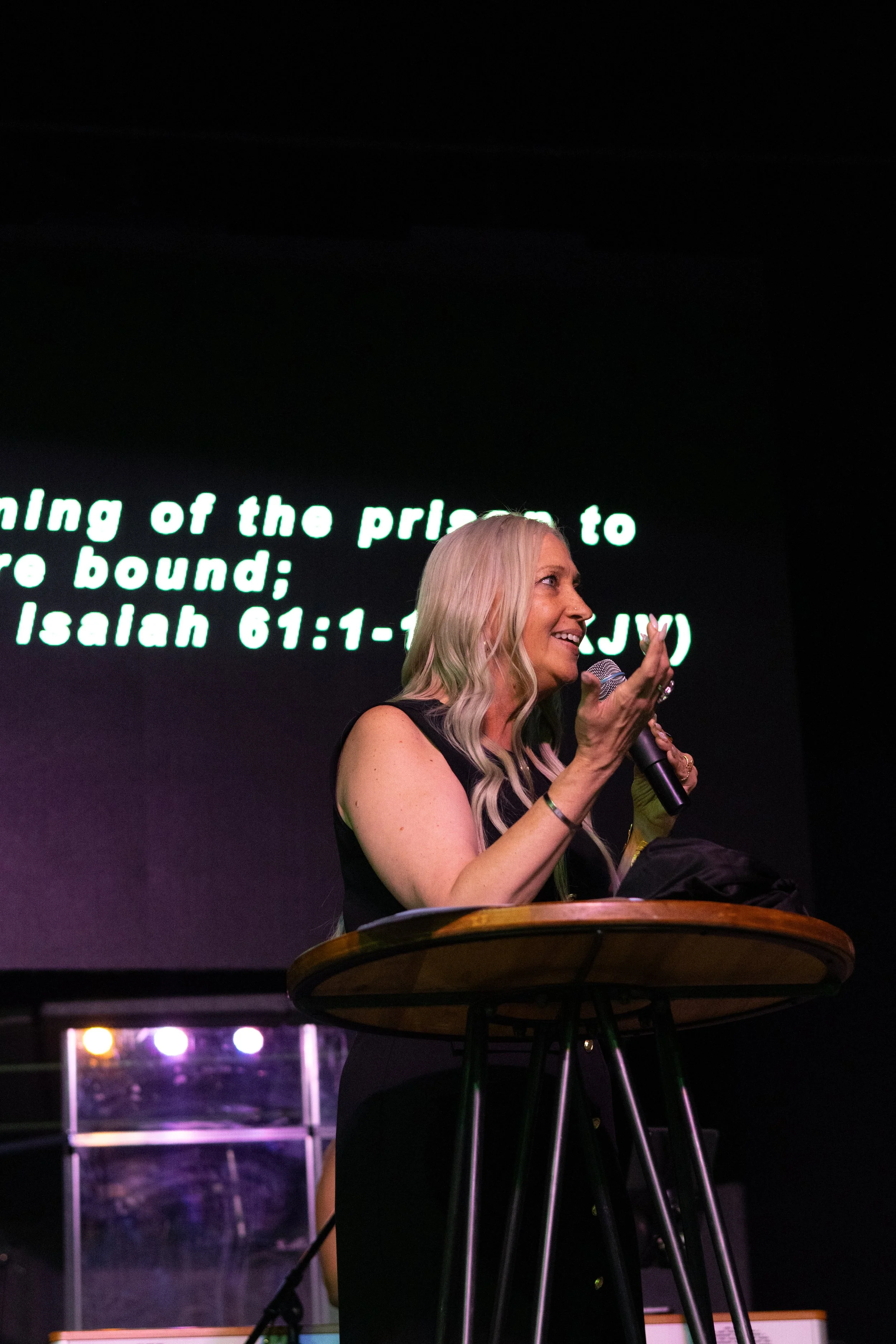 Woman with long blonde hair speaking into a microphone on stage, with a large screen displaying a biblical verse in the background.