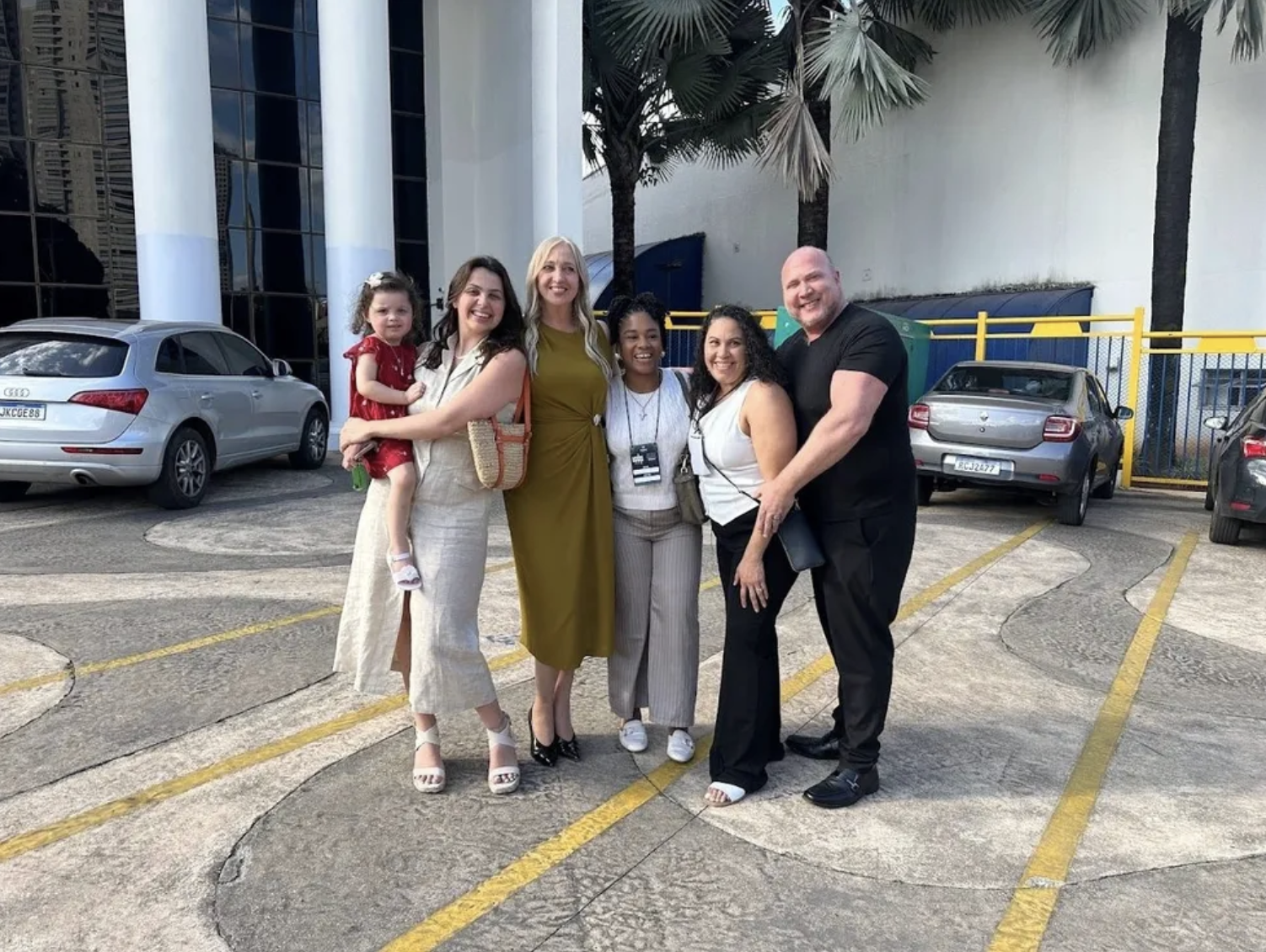 A group of six people posing together outdoors, with three women, one man, a girl, and a woman with glasses, standing on a parking lot with cars and palm trees in the background.