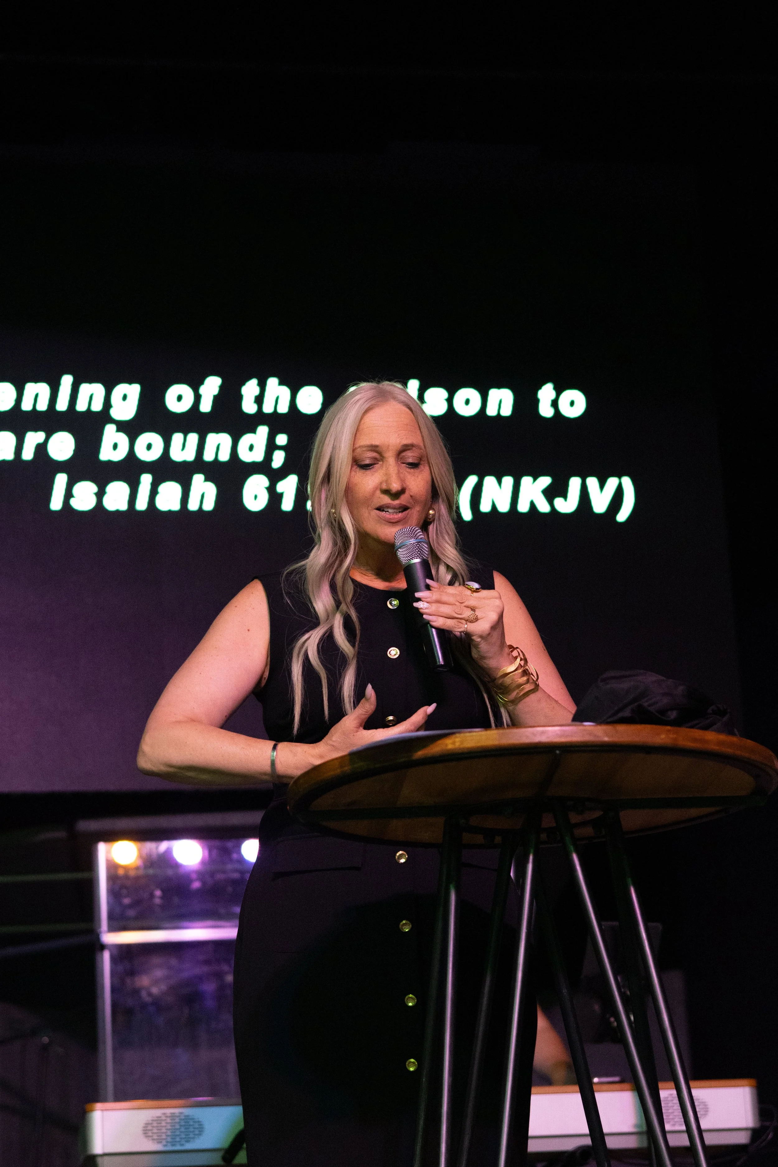 A woman with long blonde hair speaking into a microphone at a podium during a presentation or speech, with lyrics from a song displayed on a large screen behind her.