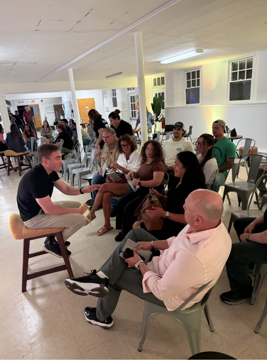 A young man in black shirt and beige pants seated on a chair, talking to a group of people sitting in chairs in a room with white walls and large windows, during an event or gathering.
