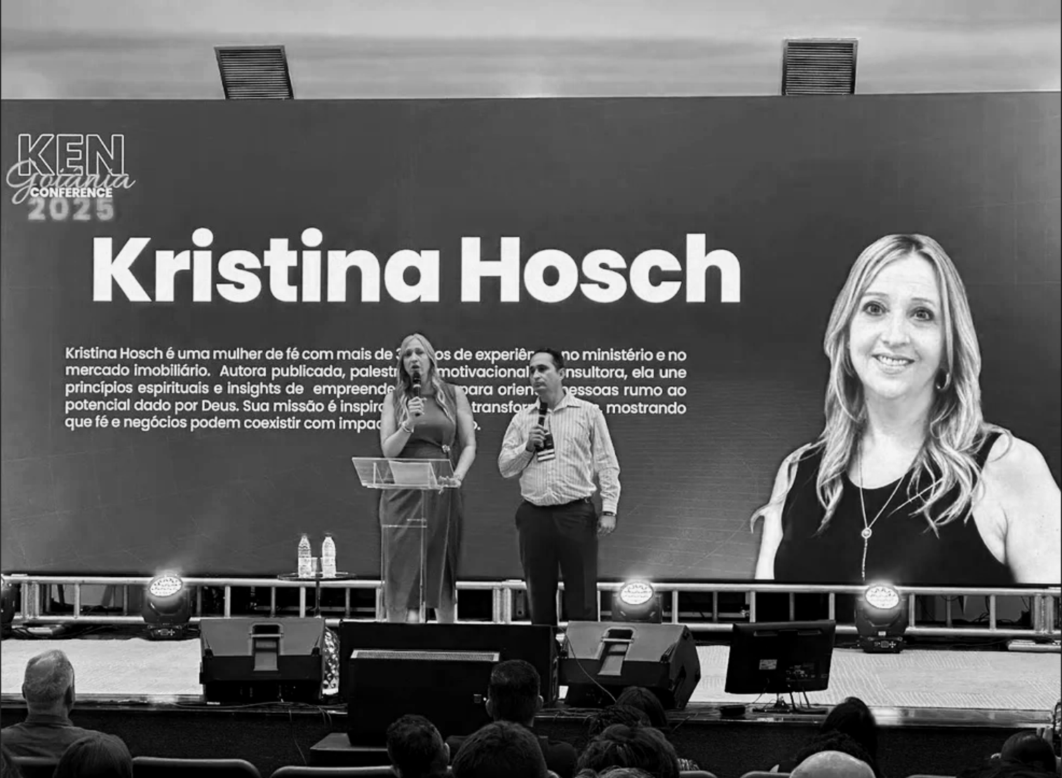 A woman and a man speaking on stage at a conference, with a large banner behind them displaying a photo of a woman and text about Kristina Hosch.