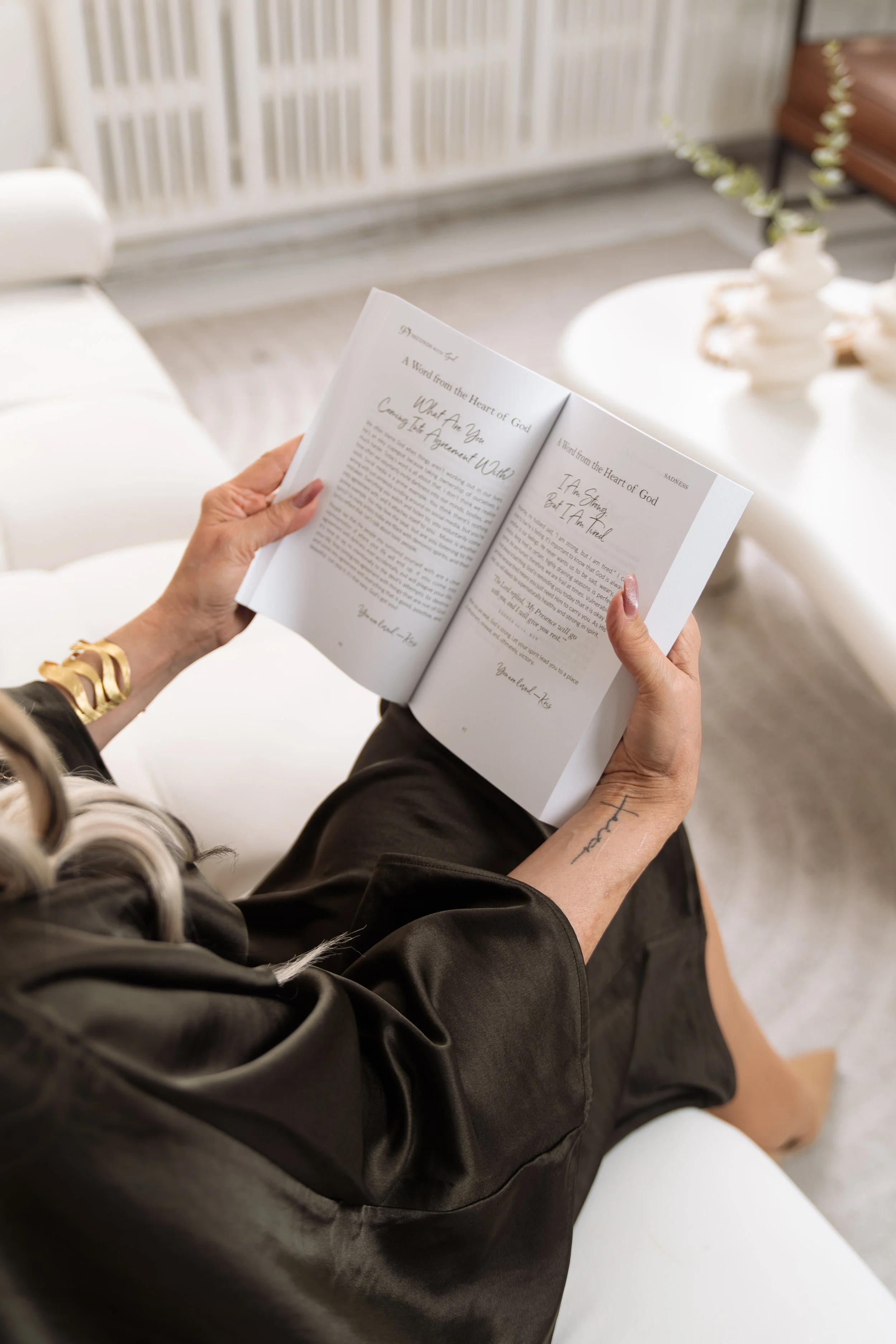 Person reading a book titled 'A Word from the Heart of God' in a cozy, well-decorated living room with white furniture and a sculptural vase with flowers.