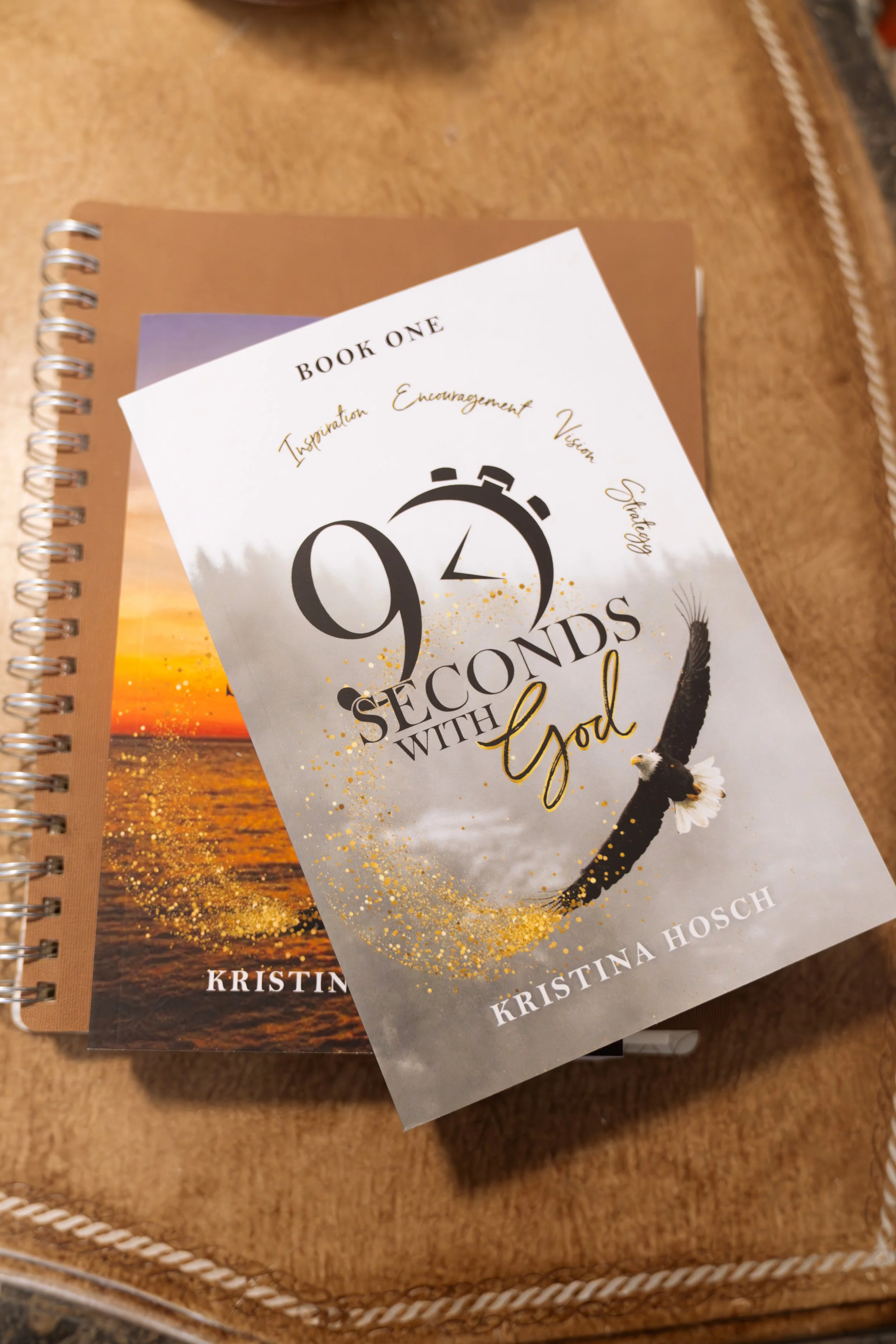 Book cover titled "90 Seconds with God" by Kristina Hosch, featuring a black eagle with outstretched wings, gold splatters, and a clock graphic, placed on a wooden table along with another book.