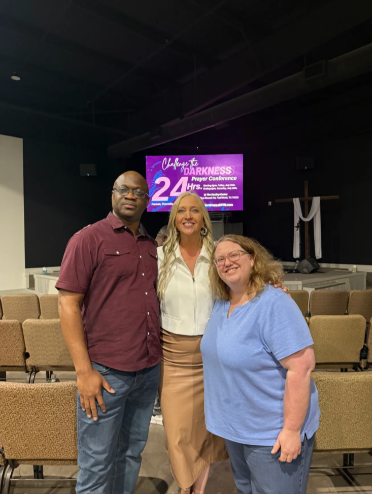 Three people standing in a church auditorium, with rows of empty chairs behind them and a digital screen displaying 'Challenge the Darkness Prayer Conference' in the background.