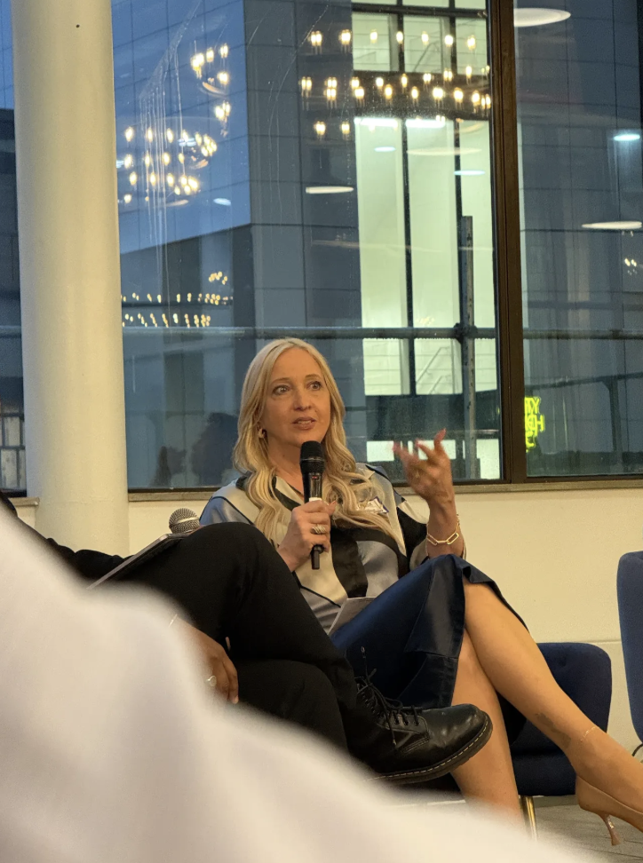 A woman with blonde hair speaking into a microphone during a panel or interview, seated on a blue chair near a large window with cityscape reflections.