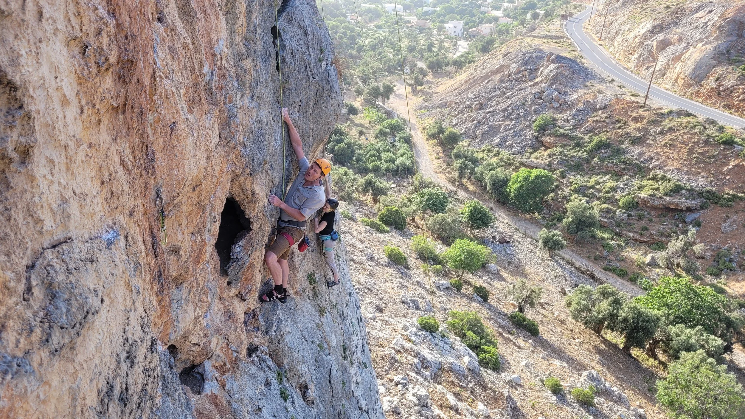 Deux personnes pratiquent l'escalade en falaise, l'une en premier plan avec un casque orange, et une autre derrière. La falaise est rocheuse, et la scène se déroule en hauteur, avec une vue sur les montagnes et une route serpentant en contrebas.
