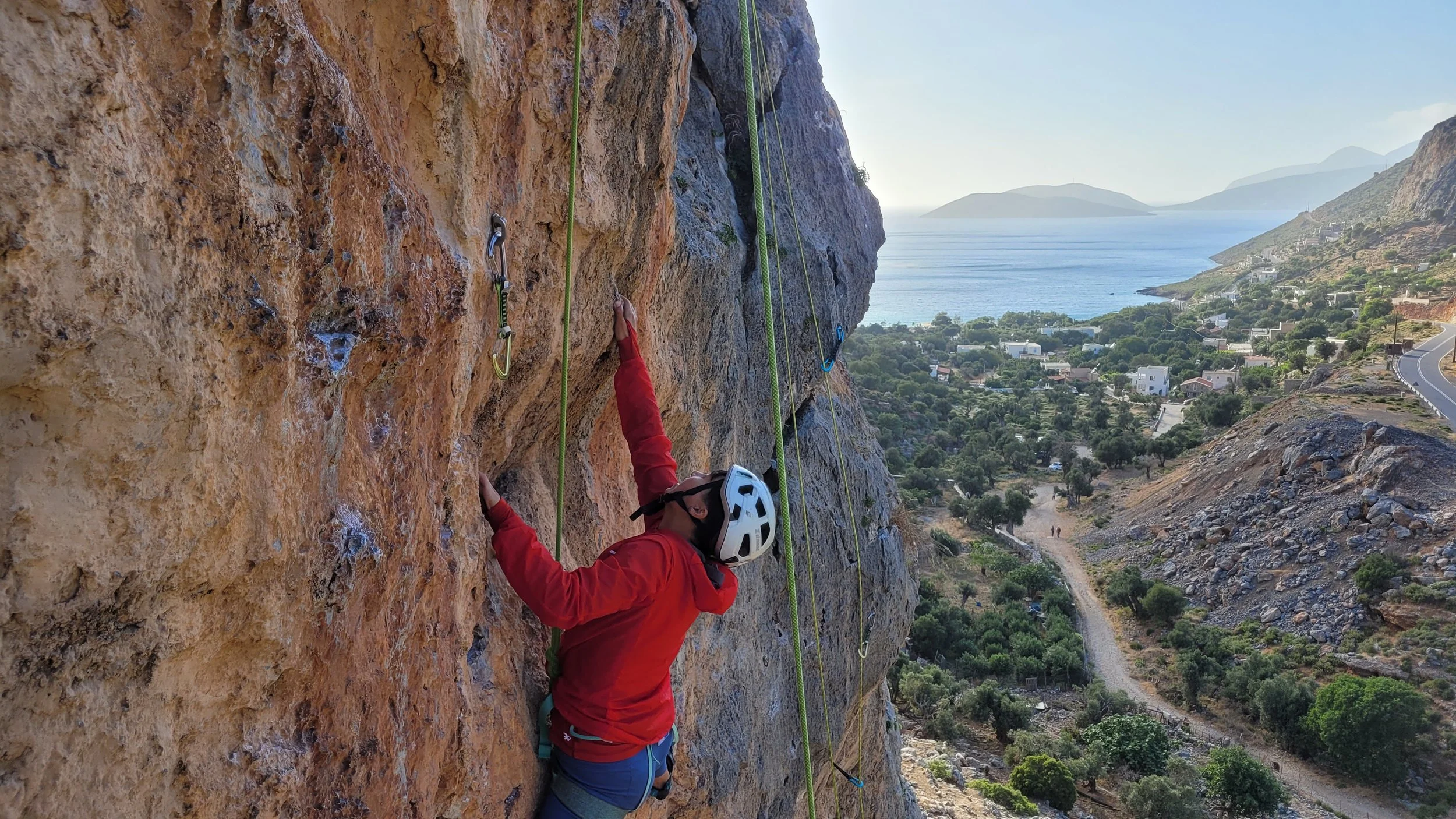 Un homme faisant de l'escalade rocheuse sur un mur en falaise, avec vue sur la mer et la côte en arrière-plan.