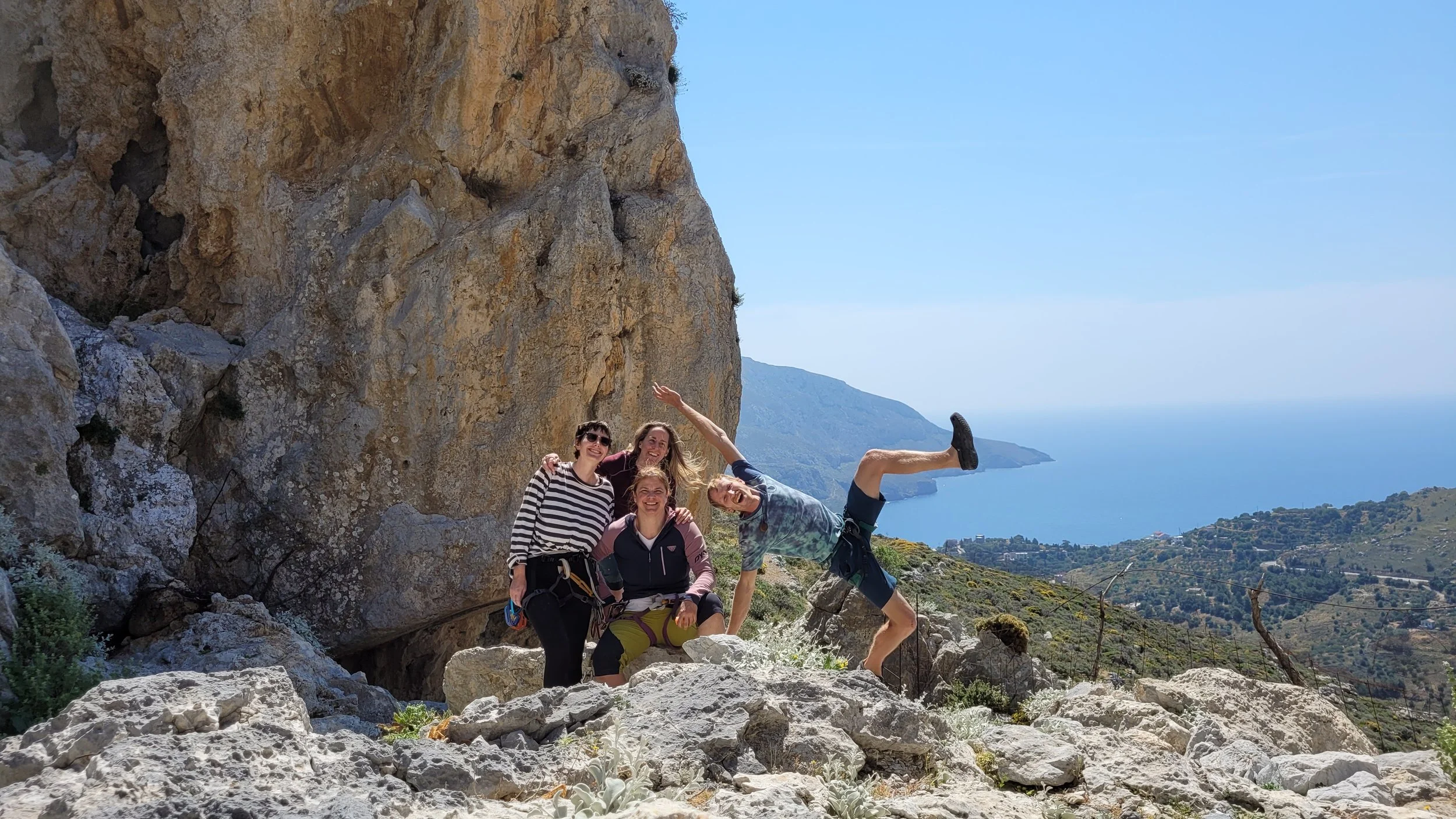 Groupe de quatre personnes en randonnée sur un paysage escarpé avec vue sur la mer et des collines en arrière-plan, devant un gros rocher rocheux.