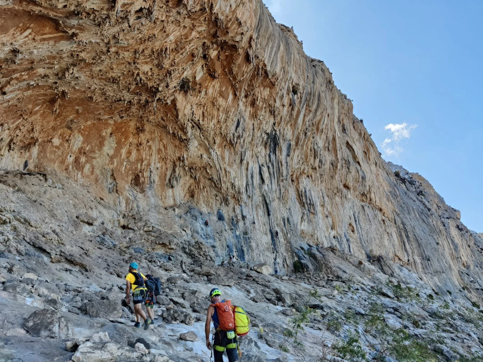 Groupe de trois alpinistes en montagne, équipés de casques et de sacs à dos, escaladant un terrain rocheux sous une grande falaise rocheuse.