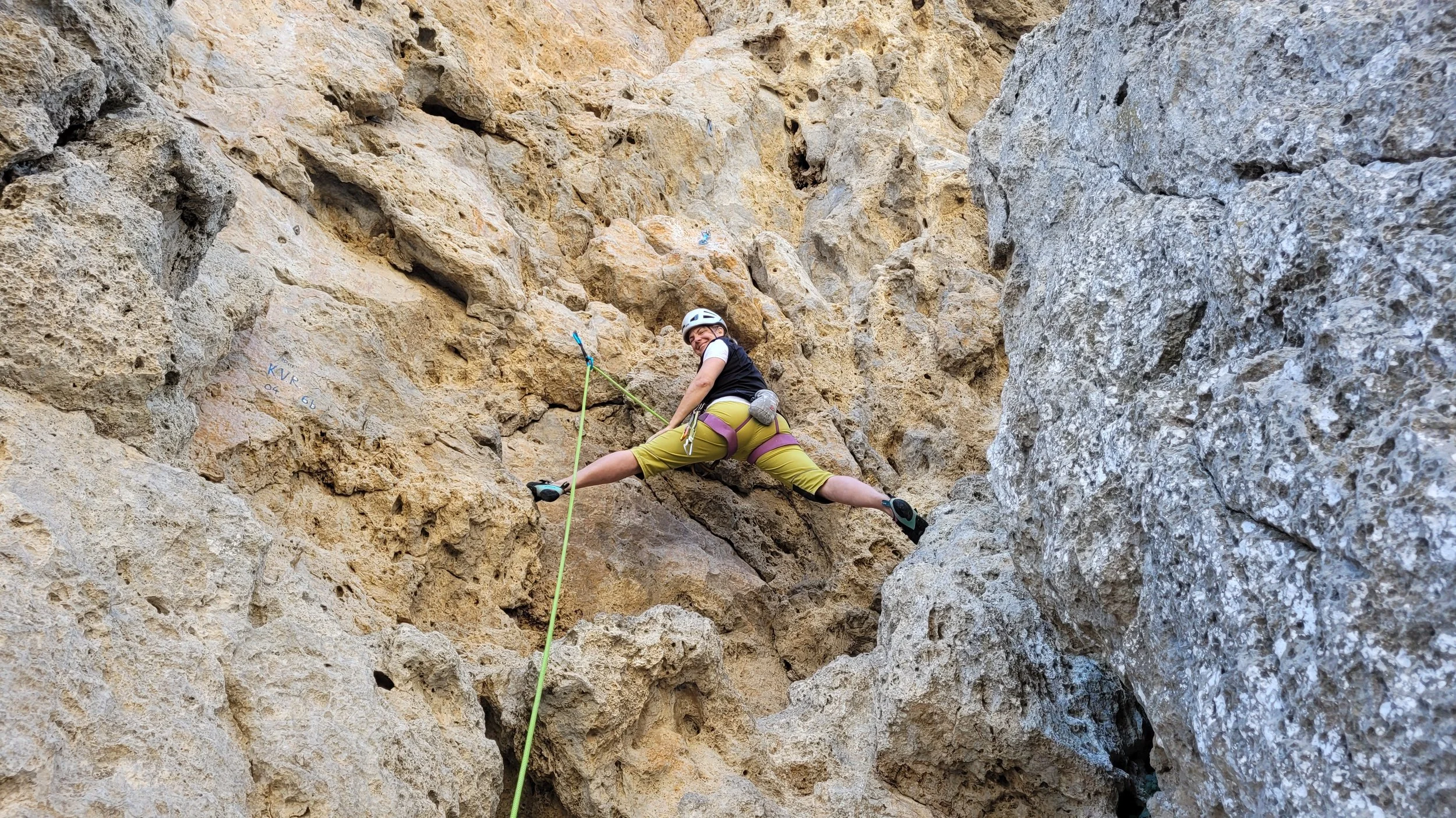 Une femme fait de l'escalade en roche dans une falaise rocheuse, portant un casque, un harnais et des chaussons d'escalade.