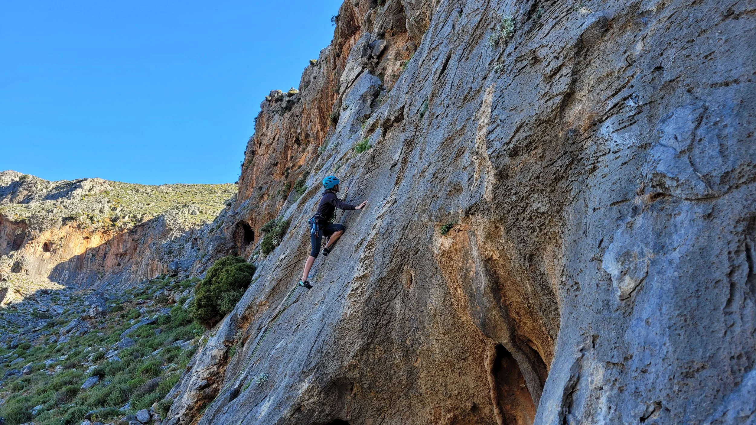 Coureur en escalade sur un rocher en plein air sous un ciel bleu. Le terrain est rocheux avec de la végétation verte autour.