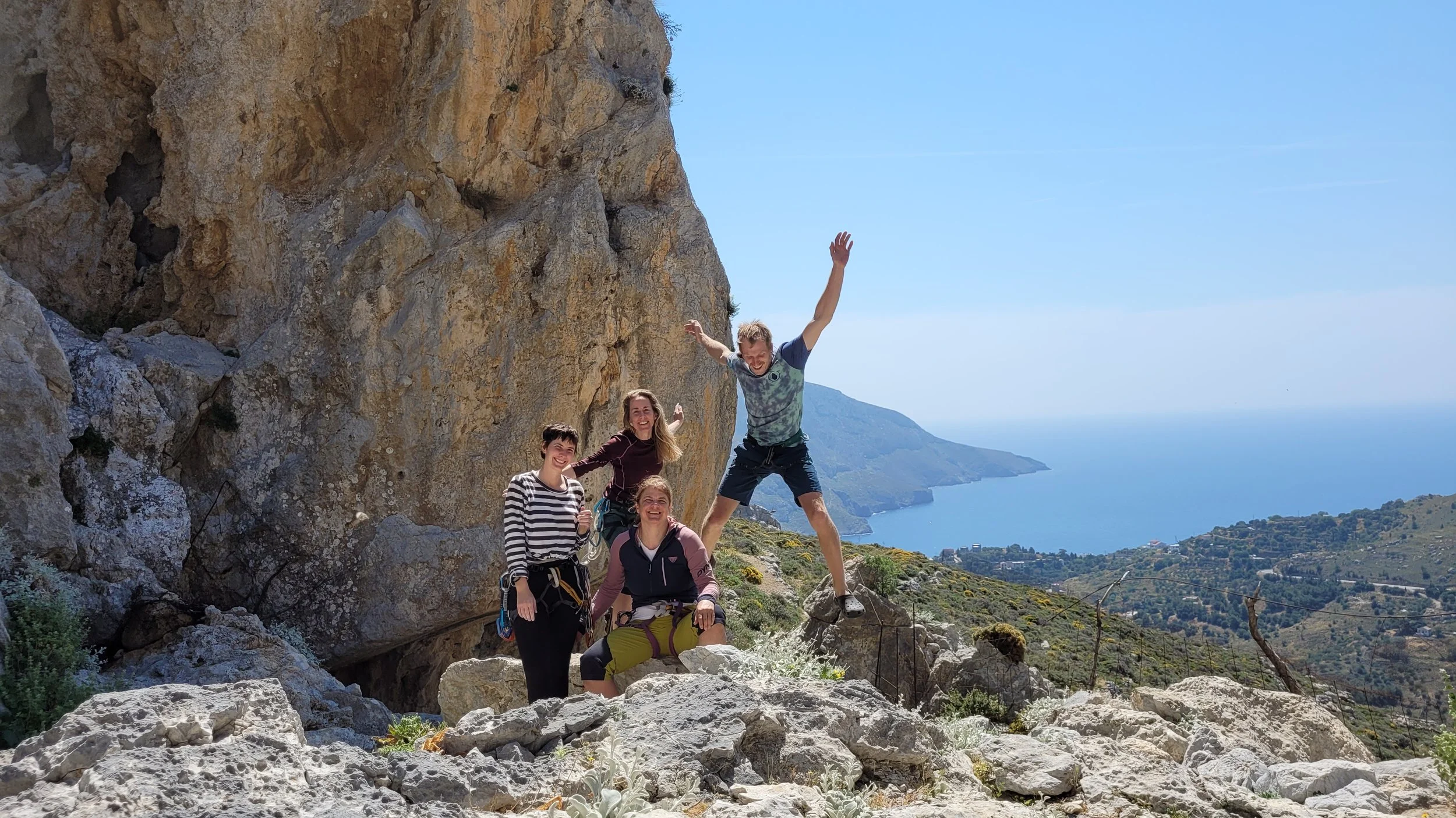 Groupe de quatre amis en randonnée de montagne sur un terrain rocheux avec vue sur la côte et la mer, le ciel clair et ensoleillé.
