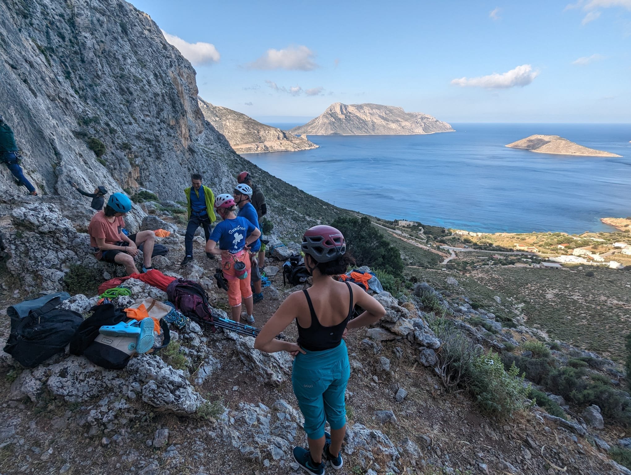 Groupe de randonneurs se reposant sur une falaise avec vue sur la mer et des îles en arrière-plan, portant des casques et du matériel d'escalade.